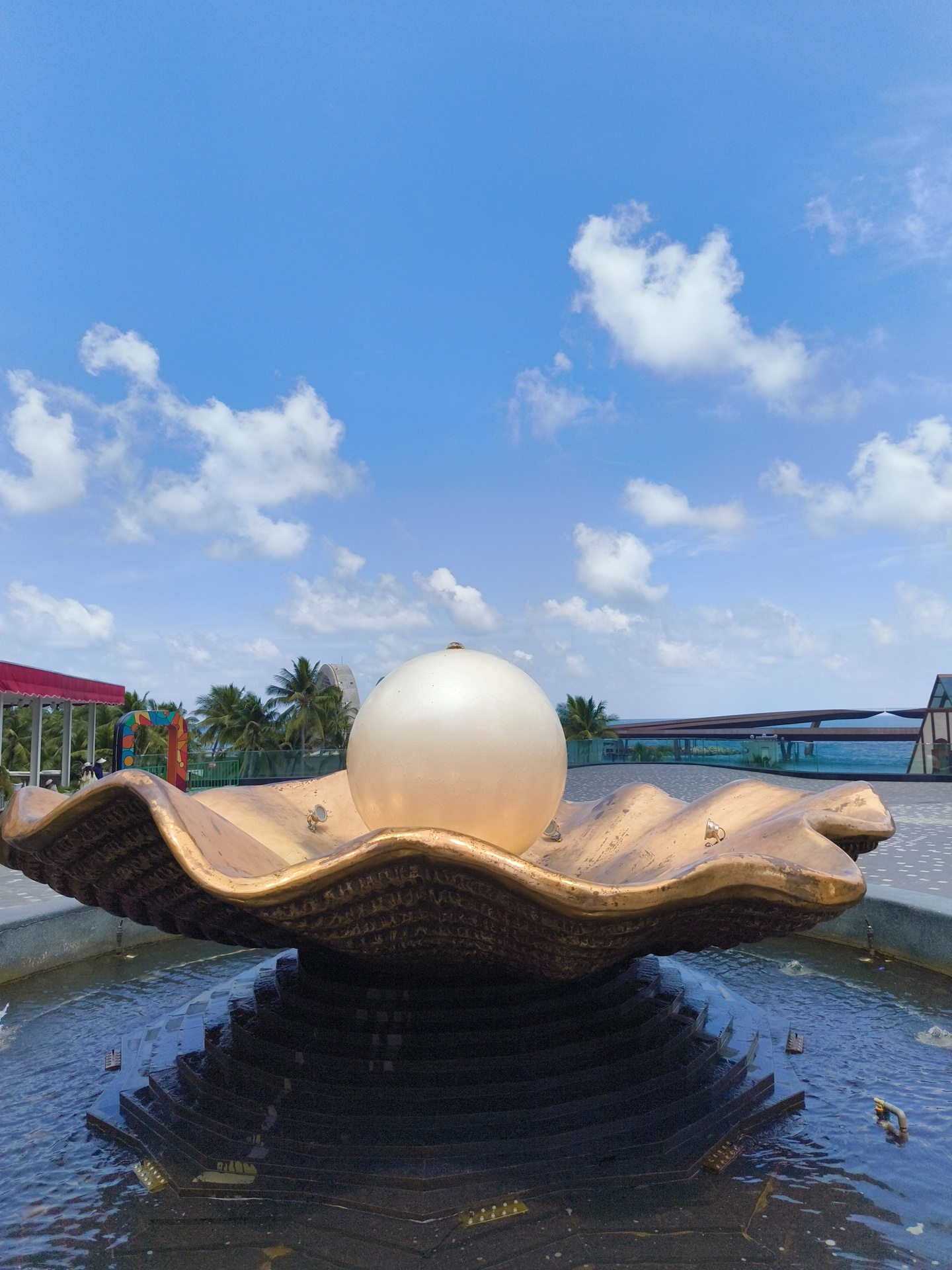 A fountain featuring a large, golden, shell-shaped basin with a luminous white sphere resembling a pearl sits at its center. The shell is textured and appears to be made of metal. Water cascades down a tiered, dark-colored base beneath the shell and into the surrounding pool, where several small water spouts are visible. The scene is set outdoors, with a clear blue sky dotted with fluffy white clouds above. In the background, palm trees and modern architectural structures are visible, including what appears to be a curving building and a bridge-like structure over water. The location is identified as Phú Quốc, Vietnam, suggesting a tropical, possibly resort or entertainment district setting. The image is captured from a low angle, emphasizing the grandeur of the fountain.