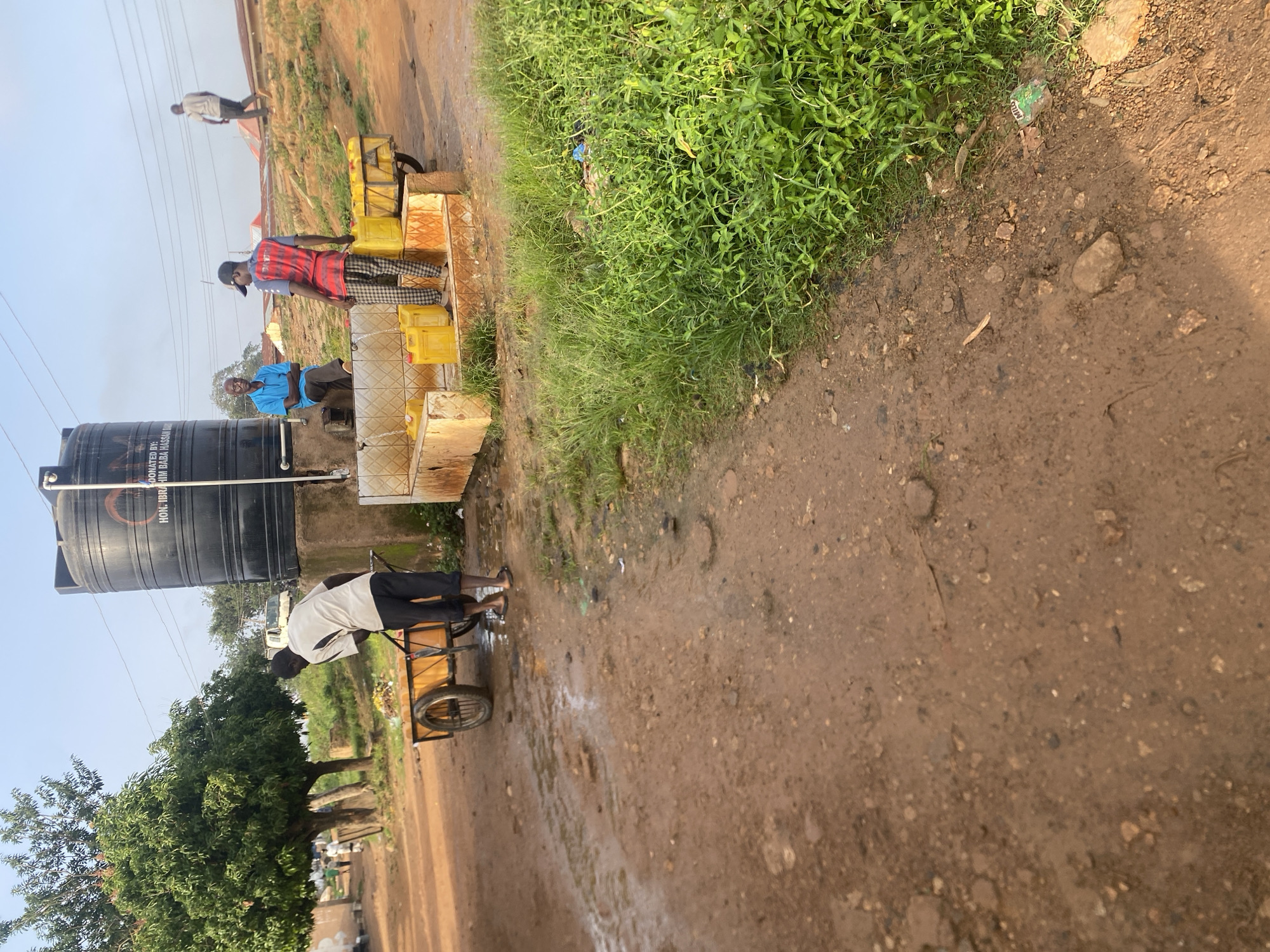 In the city of Jos, Nigeria, a man in a white shirt and black pants is pushing a two-wheeled cart laden with waste near a water dispensing station. Beside him, a man wearing a blue shirt sits on a concrete ledge, watching. Another man, dressed in a red and blue striped shirt and checkered pants, is filling yellow jerry cans with water from a tap. Behind them, a large black water tank is visible, with text that reads "DONATED BY: HON. IBRAHIM BABA HASSAN BA." In the background, several buildings with corrugated iron roofs are scattered across a sloping landscape, and another person walks away from the water station. The ground is a mix of red dirt and puddles, with green grass and vegetation growing to the right of the water area. The overall environment appears to be a semi-rural or informal settlement within Jos.