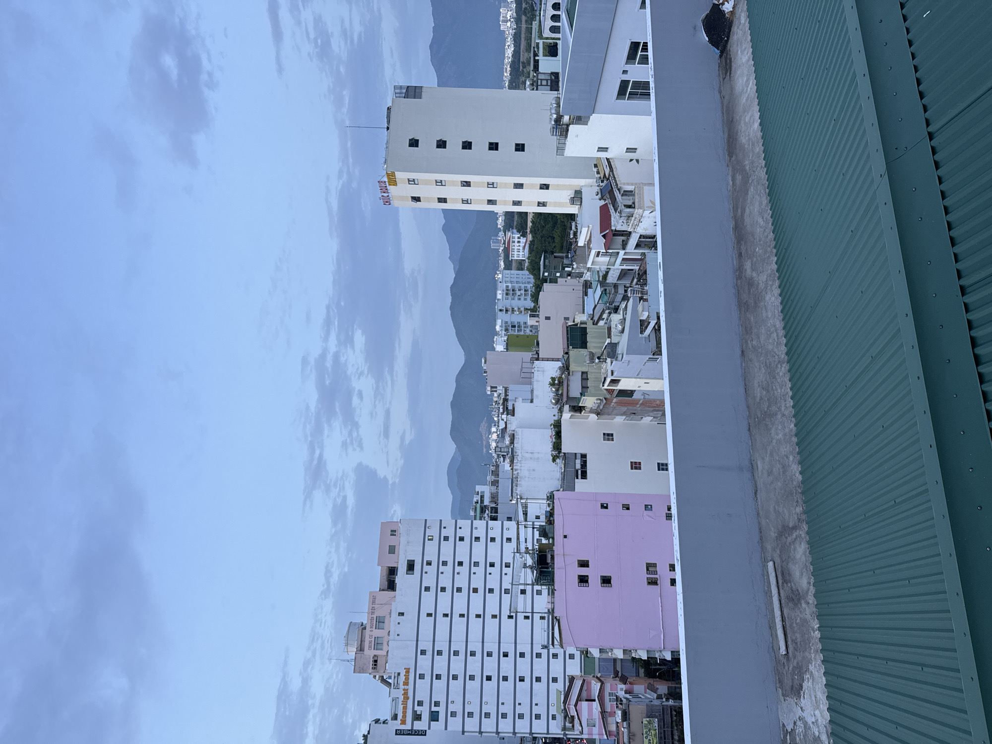 This is a photograph of a city skyline in Nha Trang, Vietnam, taken from an elevated vantage point. The image captures a collection of buildings, varying in height and color, under a cloudy sky.
In the foreground, a light grey concrete wall separates the viewer's perspective from the city below. To the right of this wall, a green corrugated metal roof occupies the bottom portion of the frame.
Dominating the mid-ground are several buildings. On the left, a tall, white building with a distinctive pattern of dark square windows is labeled "MOONLIGHT HOTEL". To its right, a narrower, taller building is white with the words "CHIC HOUSE HOTEL" in red and yellow lettering near its top. Adjacent to and in front of these, a vibrant pink building stands out. Other buildings in varying shades of white, pink, and light blue are also visible, interspersed with rooftop structures.
In the background, a range of mountains forms a silhouette against the sky, indicating a coastal or mountainous region. The sky itself is a pale blue with scattered, soft grey clouds. The overall lighting suggests either early morning or late evening. No people are visible in the frame.
