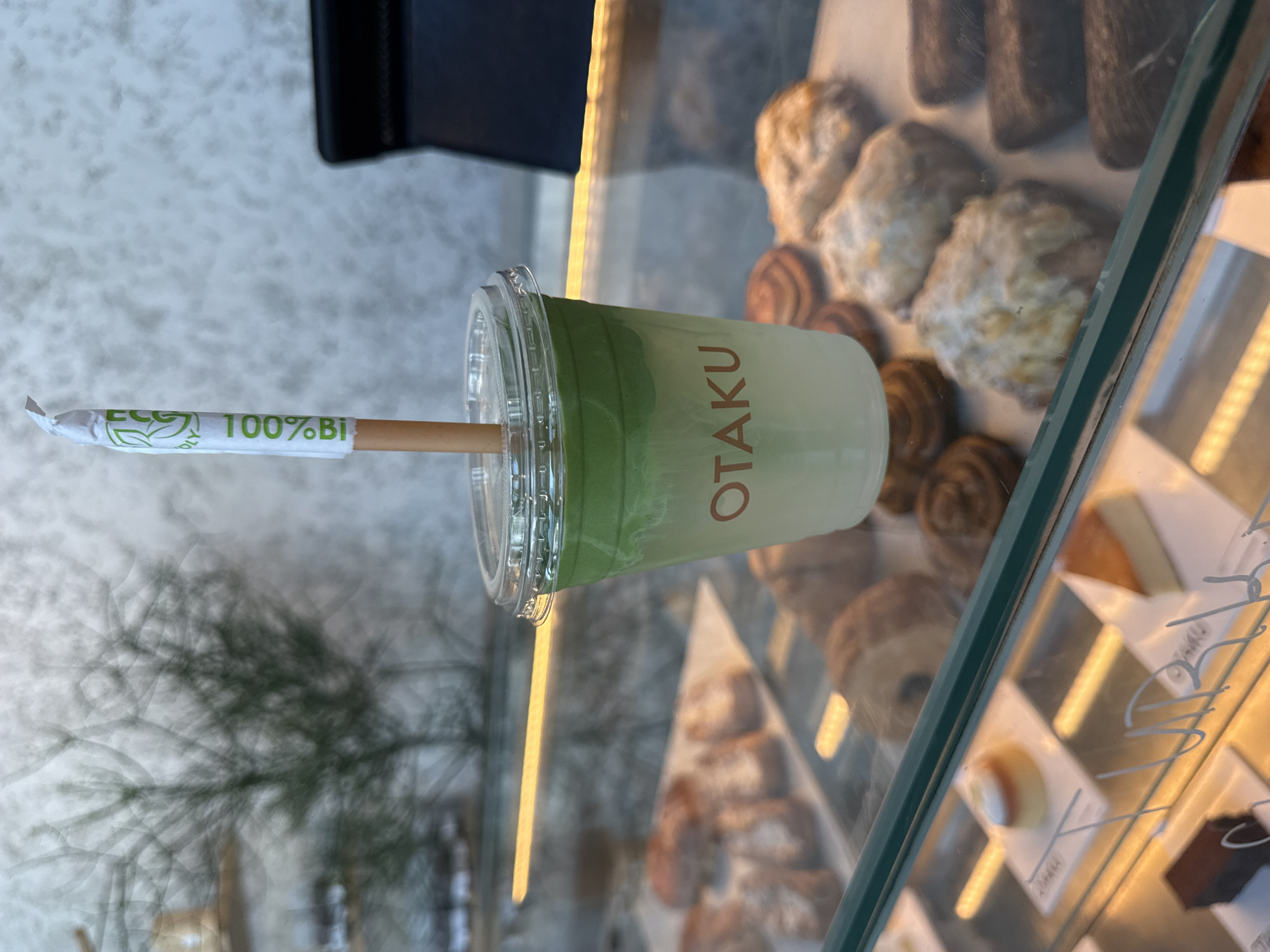 The image depicts a matcha latte in a clear plastic cup with a lid and a paper straw, resting on the glass surface of a display case. The cup has the word "OTAKU" printed on its side. The straw is wrapped in paper with "100% Bio" printed on it. Through the glass, pastries and small cakes are visible in the display case. Outside the display case, a blurred plant is visible. The setting is likely a bakery or cafe. The location is Dubai, United Arab Emirates, based on the naming of the beverage.