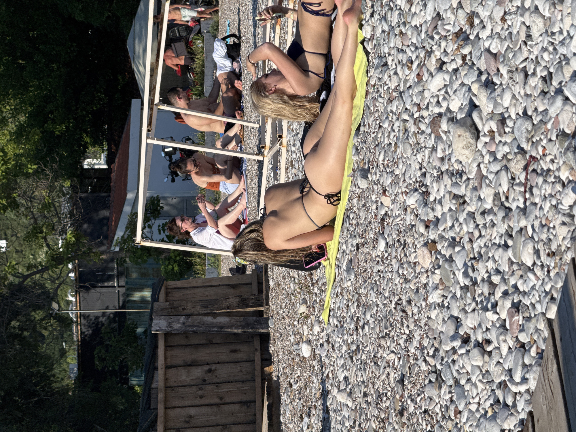 The image shows a pebble beach in Budva, Montenegro. In the foreground, two women are sunbathing on a yellow towel. The woman on the left is lying on her stomach, facing away from the camera, while the woman on the right is lying on her side, looking at her phone. In the background, several other people are visible, some sitting and some standing. They appear to be enjoying the sunny weather on the beach. The environment is typical of a Mediterranean beach, with pebbles, clear water (not visible in this frame), and surrounding vegetation. There are also some structures, possibly part of beach facilities or nearby buildings. The overall scene depicts a relaxed atmosphere on a summer day.