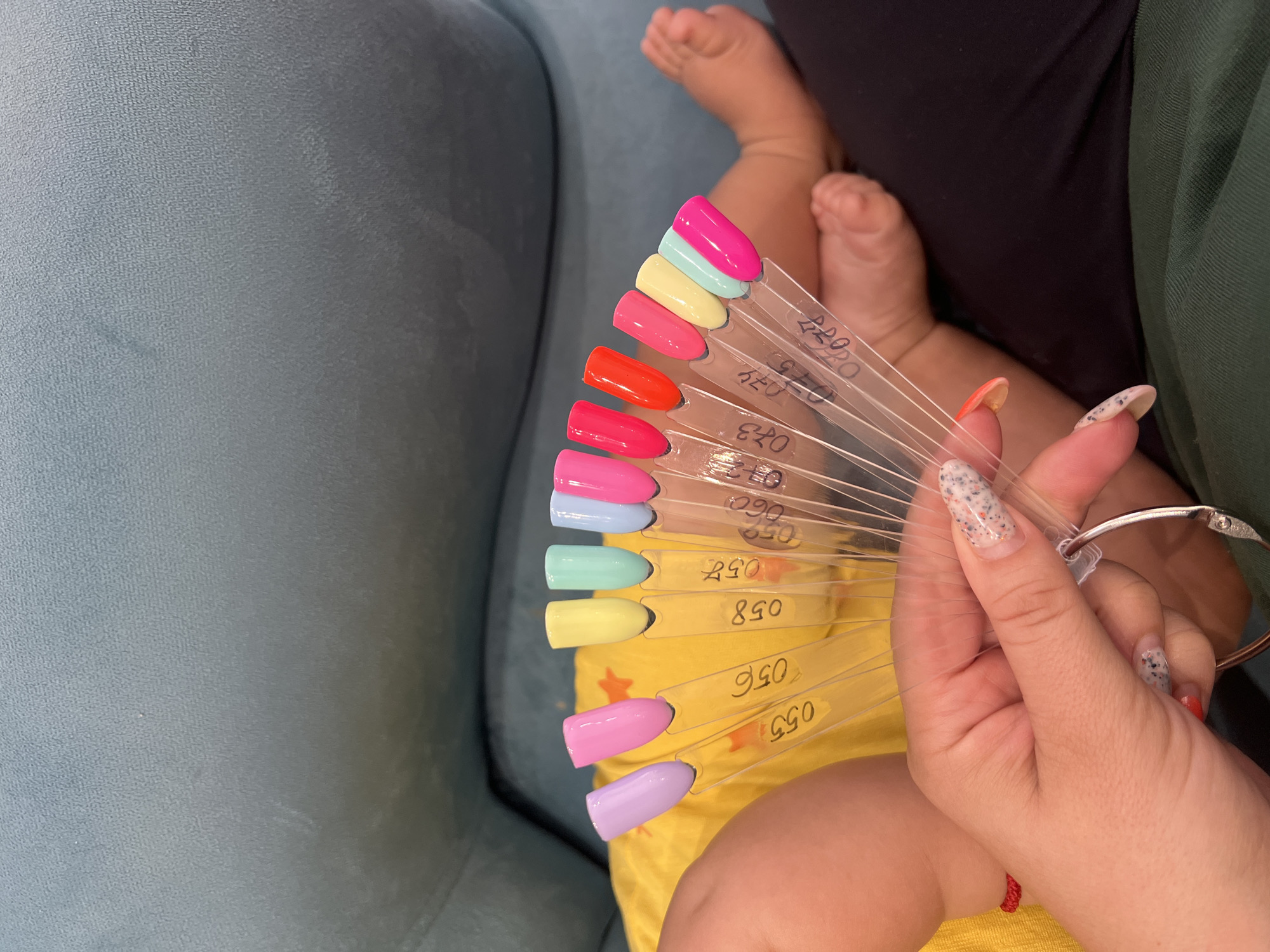 A woman's hand holds a fan of nail polish swatches against a blurred background of a blue sofa and a child's legs and feet. The swatches display various colors including lavender, pink, yellow, mint green, light blue, bright pink, red, orange, and yellow-green. Some swatches have numbers written on them, such as "055," "056," "058," "057," "059," "060," "073," "074," "075," and "077." The woman's nails are decorated with a speckled pattern. The child's legs are bare, and they are wearing a yellow garment. The environment does not provide any information about the city of Plovdiv, Bulgaria.