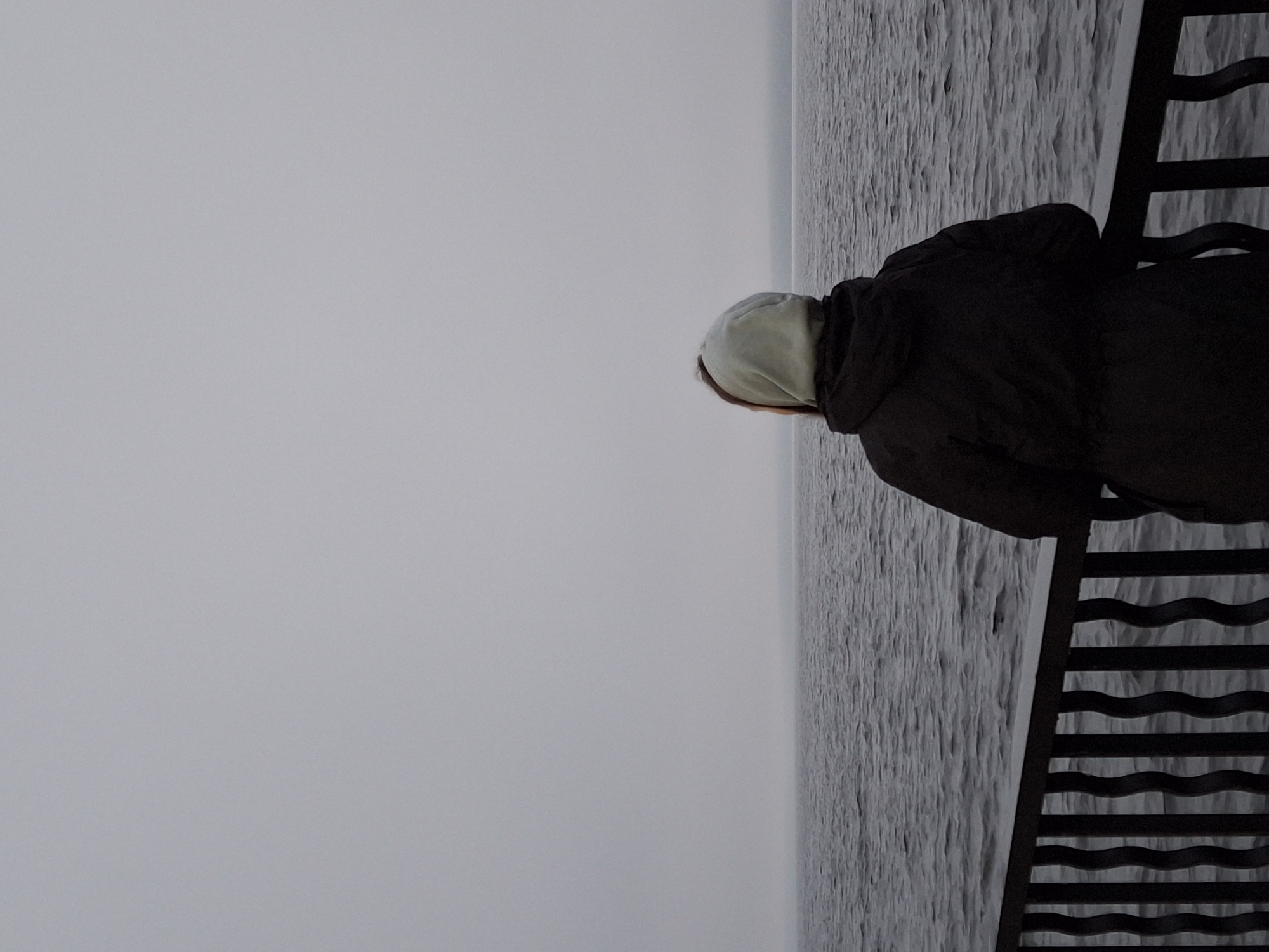 An over-the-shoulder shot captures a person from the back, wearing a dark hooded jacket and a light blue hood, standing on a dark, decorative metal railing. The person is positioned near the right edge of the frame, looking out at a vast, choppy body of water under a uniformly overcast sky. The water, a dark gray, stretches to the horizon, where a faint, indistinct landmass or shore is visible through the haze. The railing, with its ornate, wavy vertical bars, forms a distinct foreground element, framing the water. The overall atmosphere is muted and somewhat somber, indicative of a cool, possibly damp day in Chaikovsky, Russia.