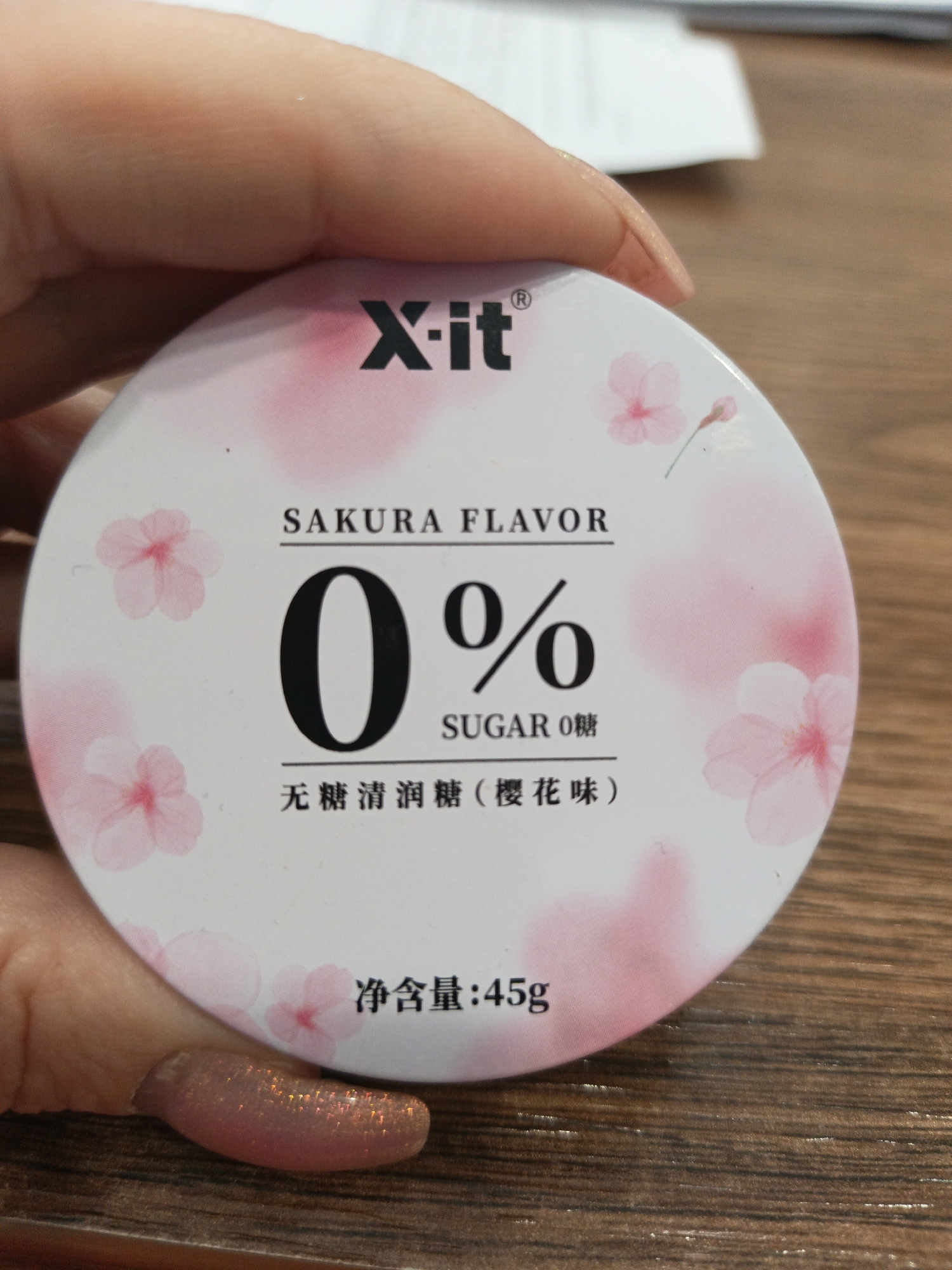 A person's hand holds a round, white container with pink cherry blossom designs. The container is labeled "X-it" and "SAKURA FLAVOR" above a large "0%" and "SUGAR 0糖". Below this is Chinese text that translates to "sugar-free moisturizing candy (cherry blossom flavor)" and "Net content: 45g". The background consists of a blurred table surface. The location is not specified and cannot be determined from this image.