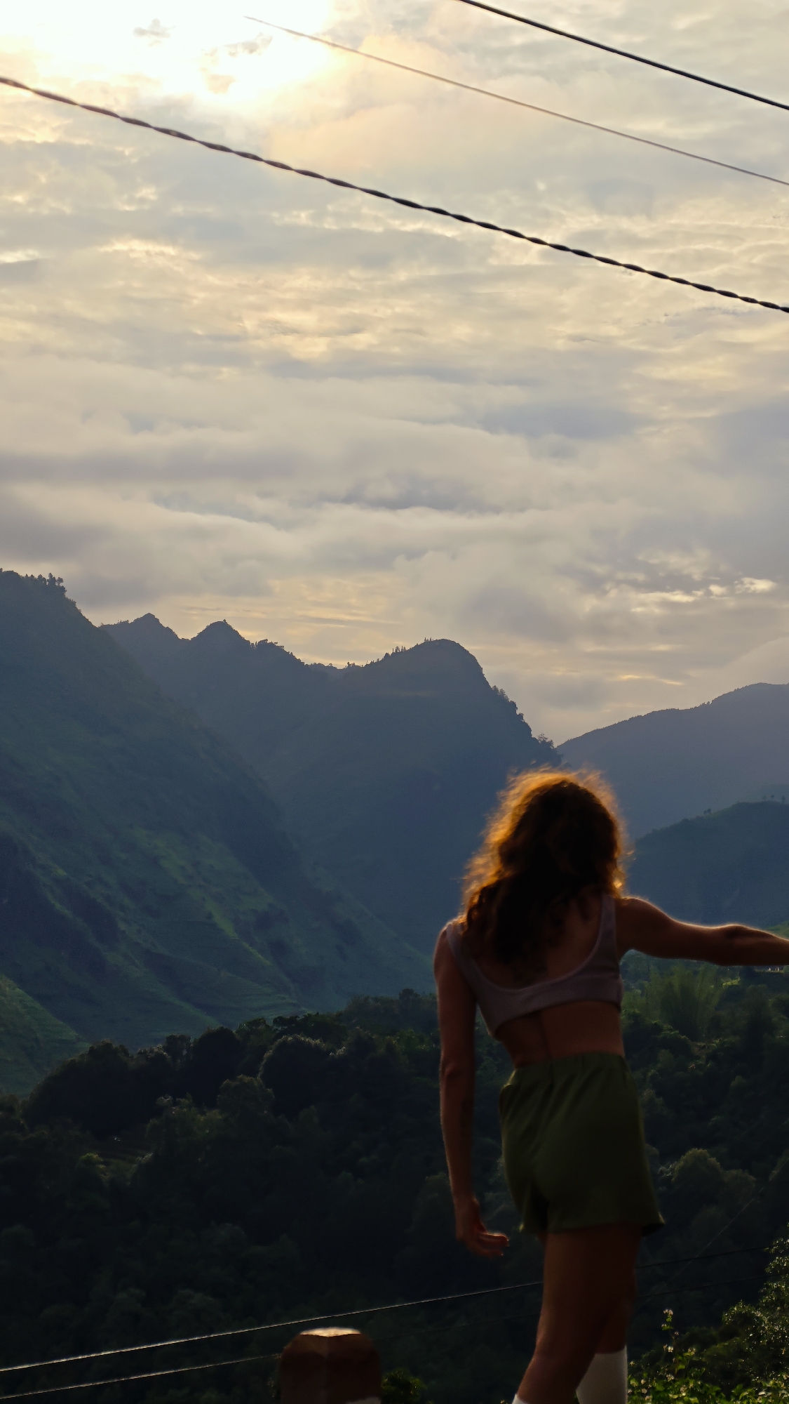 A woman in a crop top and shorts stands with her back to the camera, facing a mountainous landscape under a cloudy sky in Lũng Cú Commune, Vietnam. She is positioned on a ledge or elevated platform with posts, and her hair is illuminated by the sun. Power lines stretch across the upper part of the image. Lush green foliage covers the hillsides below the mountain range.