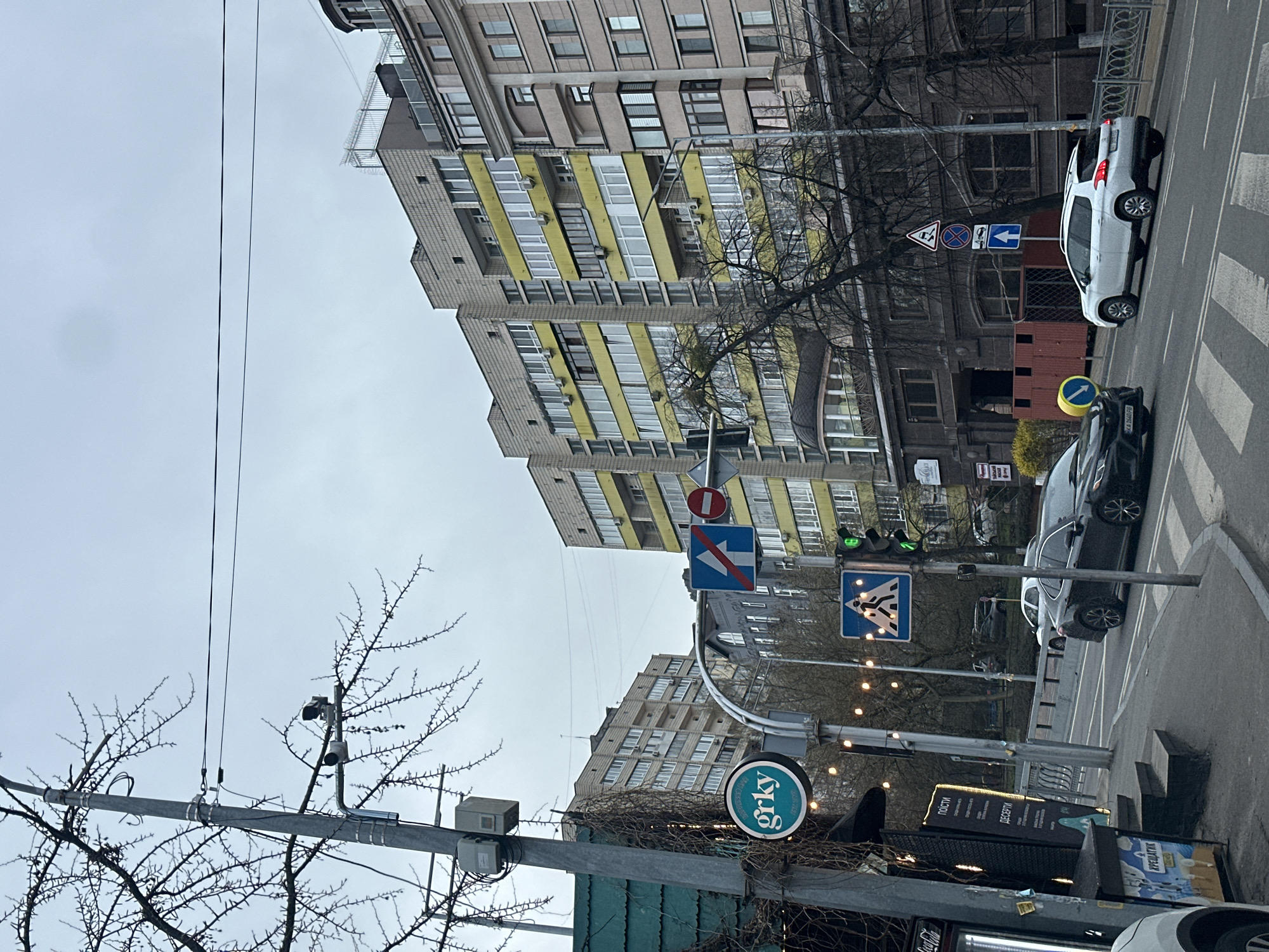 The image depicts a street scene in Kyiv, Ukraine, characterized by overcast weather. A multi-story apartment building with yellow balconies dominates the background. Street furniture includes a streetlight, signs, and wires. Two cars, one white and one black, are visible on the street. A 'grky' sign is present, potentially indicating a local business. The pedestrian crossing is marked. The scene suggests an urban environment with residential buildings and transportation infrastructure.