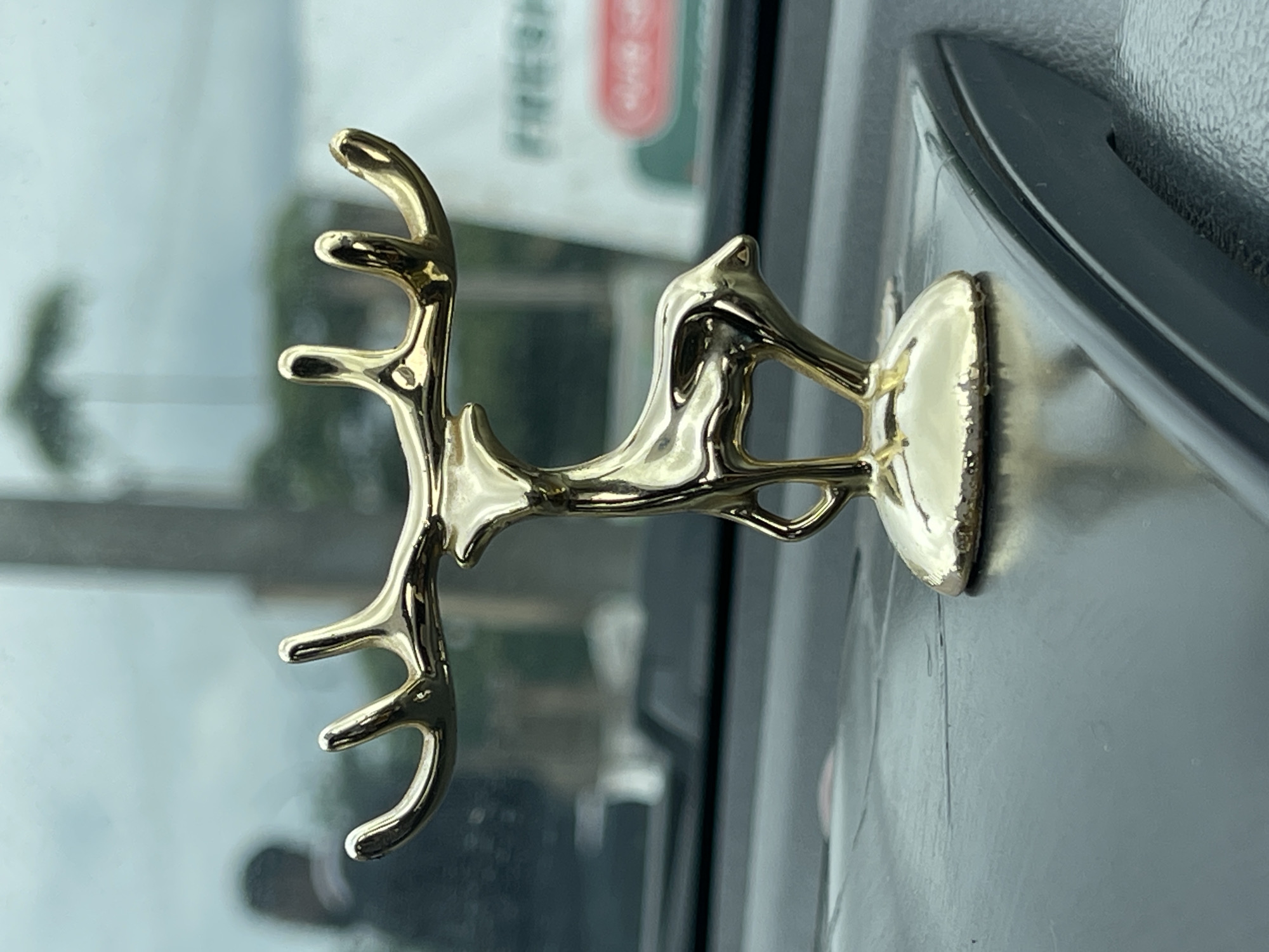 A close-up, eye-level shot showcases a small, gold-colored figurine of a deer with antlers, resting on a dark, textured surface. The figurine is the primary subject and is sharply in focus. Behind the figurine, the background is blurred, suggesting it is the interior of a vehicle. Through the windshield, a blurry figure of a person can be seen, and in the far distance, a sign with green and white lettering, possibly indicating a business, is visible. The overall lighting suggests an overcast day. This scene is located in Galadimawa Village, Nigeria.