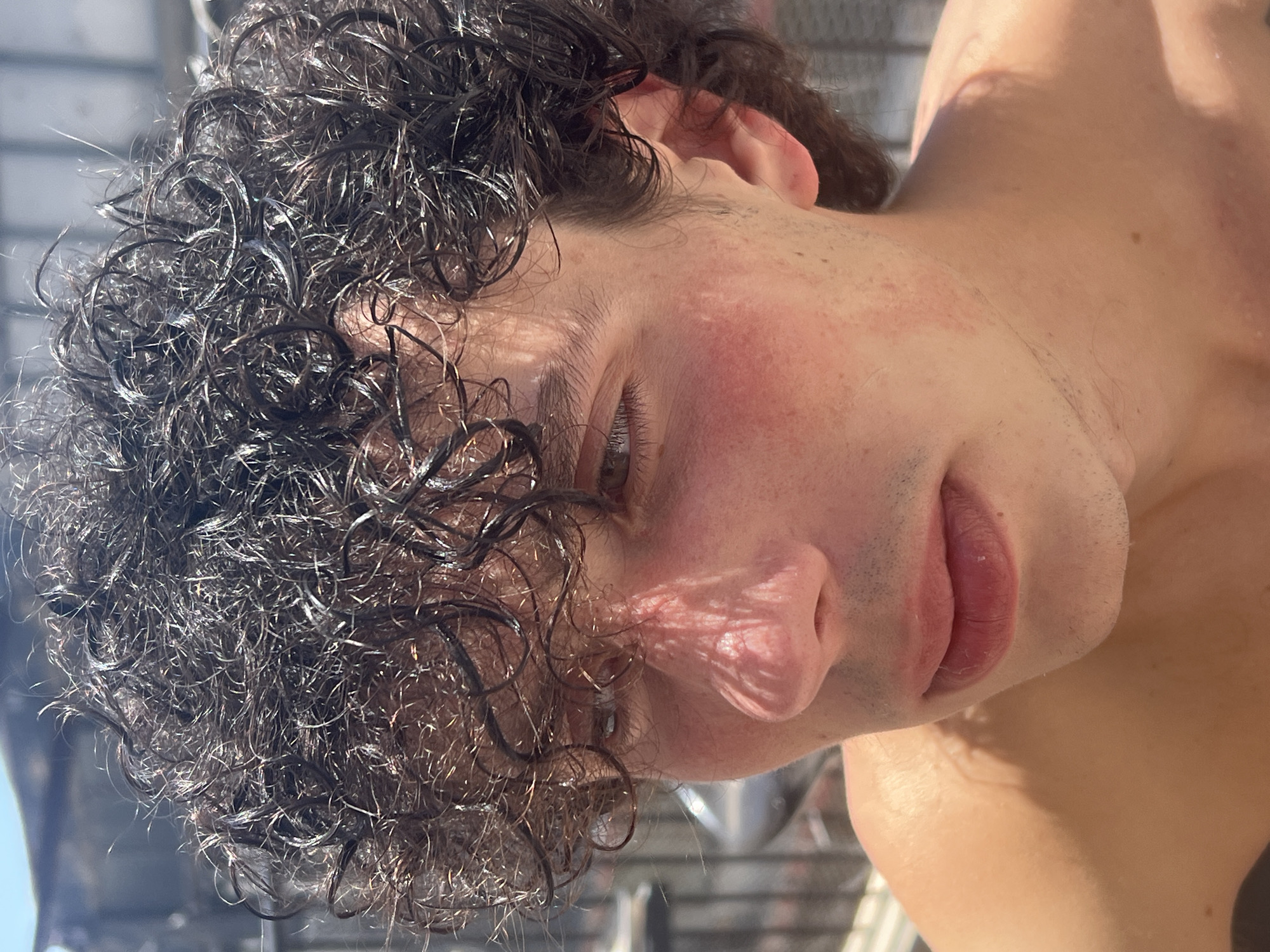 This is a close-up, medium shot of a young man with curly, dark, wet hair. He is outdoors, likely in Tel-Aviv, Israel, as evidenced by the bright sunlight and possibly a glimpse of a building or fence in the background. His face is illuminated by harsh sunlight, casting a distinct shadow pattern on his nose and cheek. He appears to have a light complexion with some freckles and a slight reddish flush on his cheeks, potentially from sun exposure. His lips are slightly parted, and his eyes are looking off to the side. He is shirtless, with his shoulder and upper chest visible. The image captures a moment of casual outdoor activity, possibly by a pool or at the beach.