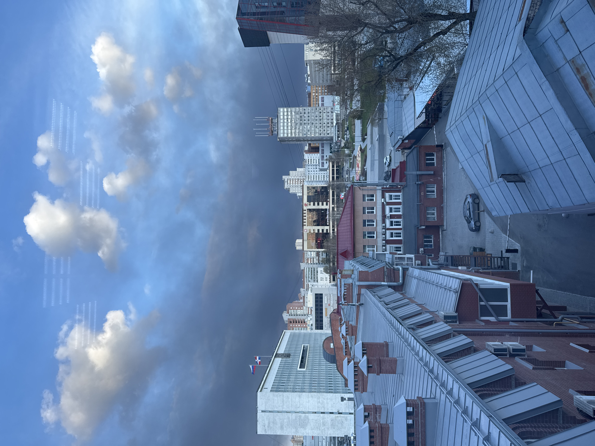 The image presents a panoramic view of an urban landscape in Perm, Russia, captured from an elevated vantage point. The scene is characterized by a dramatic sky, with clear blue patches and fluffy white cumulus clouds on the left side, transitioning to heavy, dark grey clouds on the right, suggesting a dynamic weather pattern, possibly late afternoon or early evening.
In the foreground, several buildings with traditional red brick facades and distinctive grey, ridged metal roofs are prominent. These roofs feature numerous chimneys, dormer windows, and ventilation units. Further into the mid-ground, the architecture shifts to include taller, more modern structures. A notable white or light-colored high-rise building with numerous windows stands out, topped with what appears to be an antenna array or structural elements. On the roof of another building in the mid-ground, a flagpole displays two flags; one is clearly the Russian national flag (white, blue, and red horizontal stripes), and another flag is alongside it. Additional modern high-rise buildings, including a dark-colored one with red accents, populate the background cityscape.
Street scenes are visible between the buildings, with a few parked vehicles, including a dark sedan and a smaller white car. Bare-branched trees are scattered throughout, particularly on the left side of the frame, indicating early spring or late autumn.
A peculiar detail is the presence of distinct patterns of small, rectangular, luminous reflections visible on the blue sky and cloud areas on the left side of the image. These reflections, arranged in vertical rows, suggest the photograph was taken through a glass window, possibly reflecting interior lights or architectural elements. While various signs and markings are visible on the buildings, no text is clearly legible due to distance and resolution.