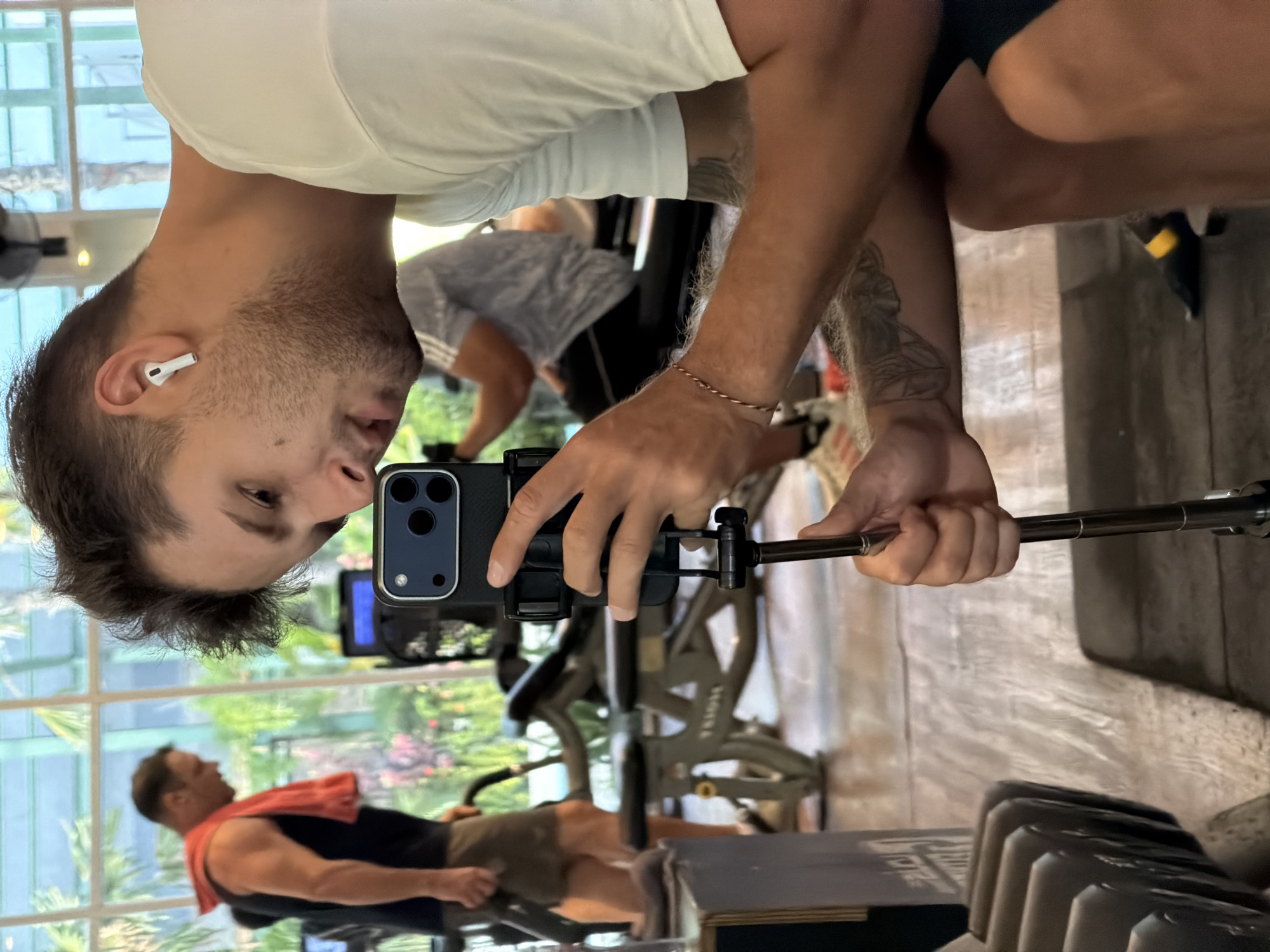 The image captures a man in a modern fitness center, likely in Denpasar Barat, Indonesia, actively engaged in taking a selfie or recording a video during what appears to be a workout. The original photograph is oriented sideways (rotated 90 degrees clockwise).
In the foreground, the man, who has short brown hair and light stubble on his face, is wearing a plain white t-shirt and dark shorts. A white Apple AirPod is visible in his right ear. He holds a black smartphone, mounted on an extending grip or selfie stick, with both hands, focusing intently on the screen. Tattoos adorn both of his forearms, and a thin, copper-colored bracelet is worn on his right wrist.
The background reveals a spacious gym environment with various exercise machines, including a stationary bike. Large, clear windows stretch across the back, allowing ample natural light to flood the room and showcasing a vibrant outdoor scene of green trees and tropical foliage. In the mid-ground, another man is visible, possibly on a different piece of equipment, with a red towel draped over his shoulders and wearing shorts with a visible Adidas logo (seen in reflection). A rack of metal dumbbells is partially in view in the lower right corner, further confirming the gym setting. The bright lighting suggests it is daytime.
