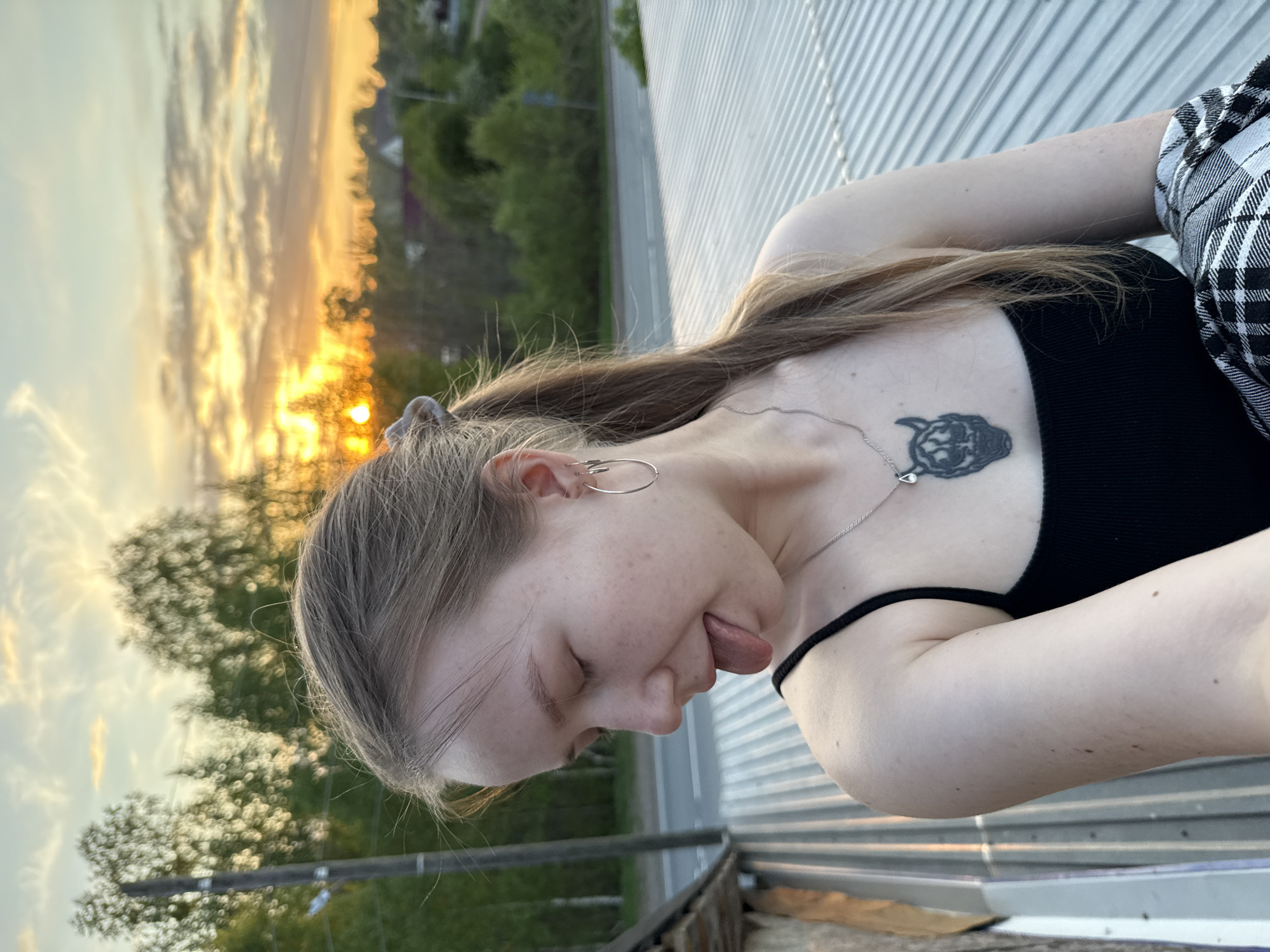 A young woman is seen close-up from a slightly elevated position, seemingly standing on a corrugated metal surface. She is wearing a black tank top and a black and white plaid shirt. A silver necklace and hoop earrings are visible. She has a tattoo of a cat silhouette on her upper chest. Her tongue is sticking out. The background reveals trees and a setting sun, suggesting it's dusk. The location is unspecified, and no information indicates the city is Unknown, Russia.