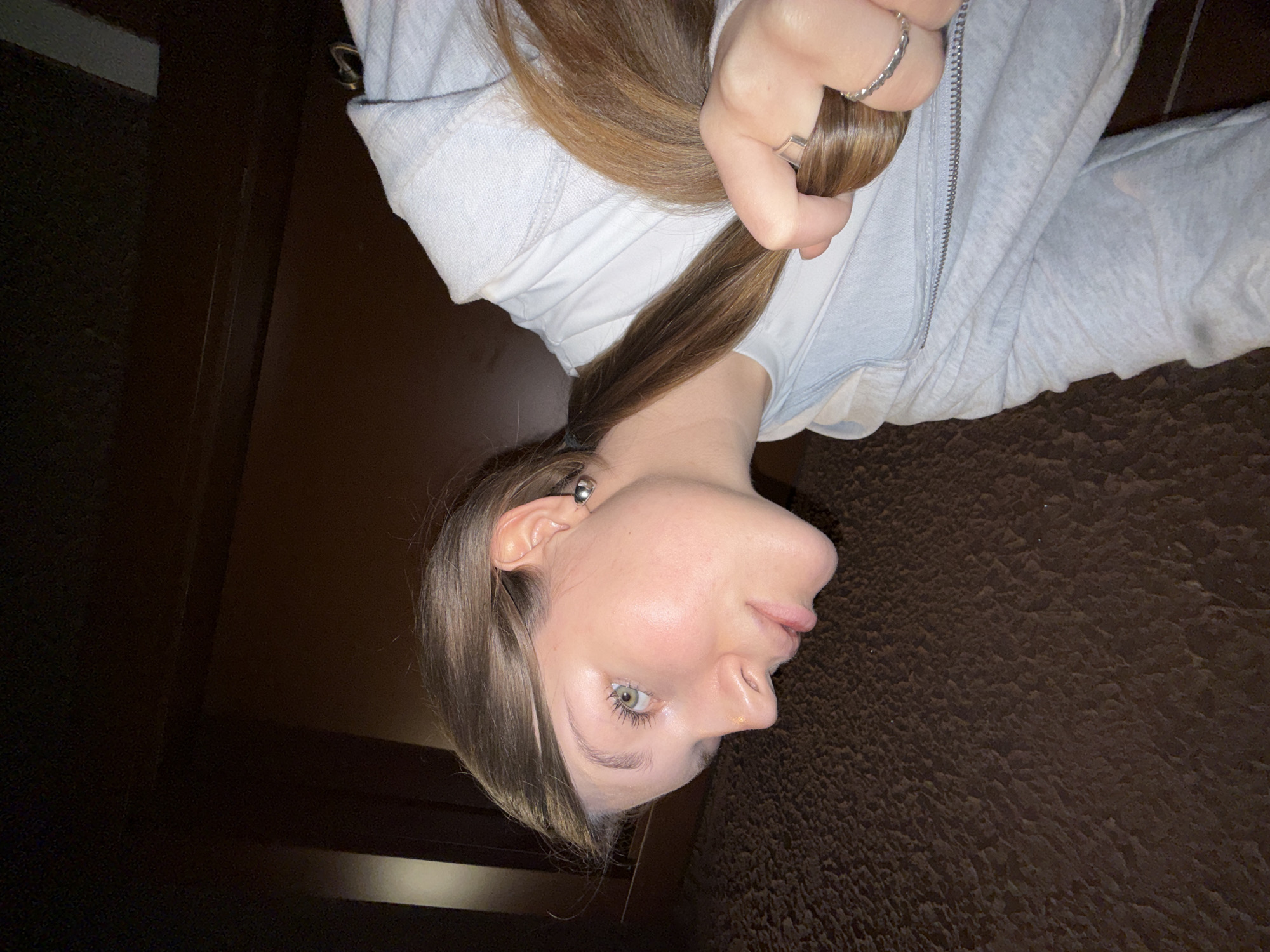 A young, light-skinned woman is depicted in the image. She has shoulder-length brown hair with lighter highlights, fair skin, and a noticeable nose. She is wearing a light gray hoodie with a zipper. Her hair is partially held in her hand. She has a ring on her finger. The background consists of a dark brown carpet or rug and a dark brown wooden door. The location is not discernible from the image.