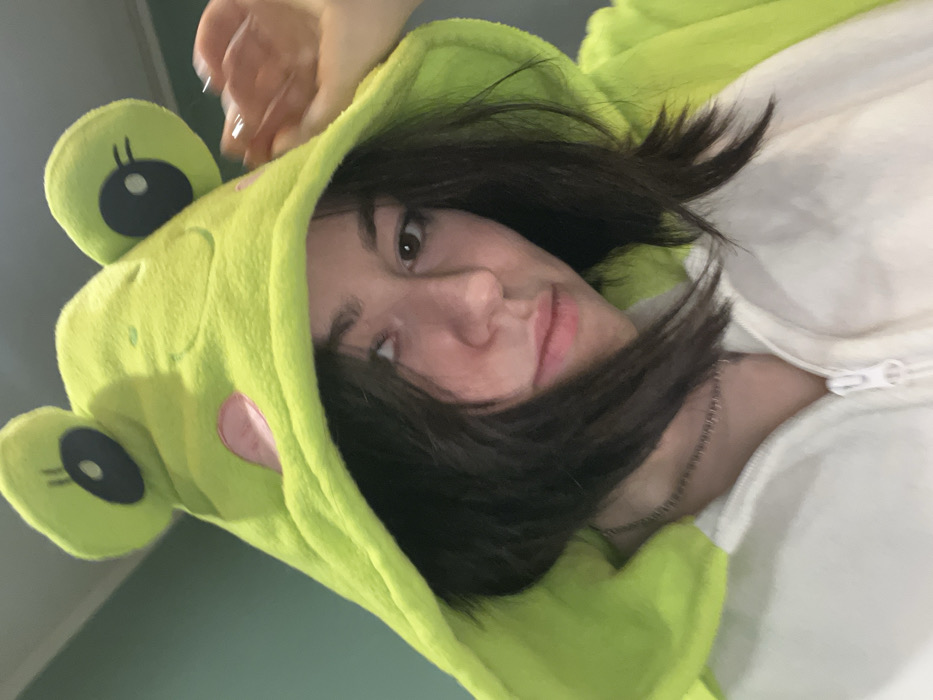 A young woman poses, partially obscured by a green frog costume hood featuring prominent eyes and a pink cheek. The hood frames her face, with dark brown hair falling across her forehead and around her shoulders. Her skin appears fair. She is wearing a white zippered garment. A delicate silver chain is visible around her neck. The photo is taken from a low angle.