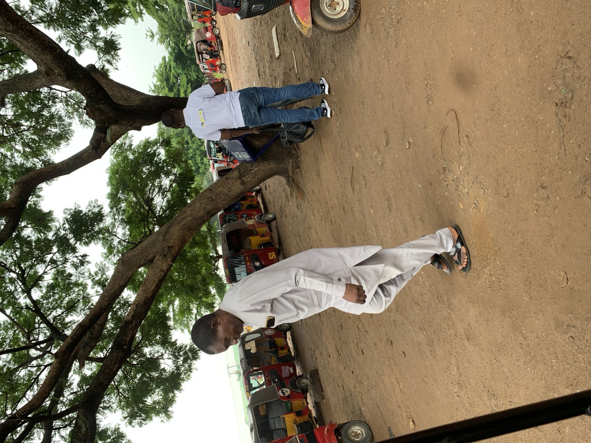 Two men are in an outdoor, dusty lot with several red and yellow auto rickshaws parked around them. A large tree with spreading branches provides shade over the scene. The man in the foreground, on the left, is wearing a light gray traditional outfit and black sandals. He is walking and looking down. The man in the background, on the right, is wearing a white polo shirt with a logo and blue jeans. He is standing and holding a dark bag. The auto rickshaws appear to be a form of local transportation. The environment suggests a transportation hub or waiting area in Abuja, Nigeria.