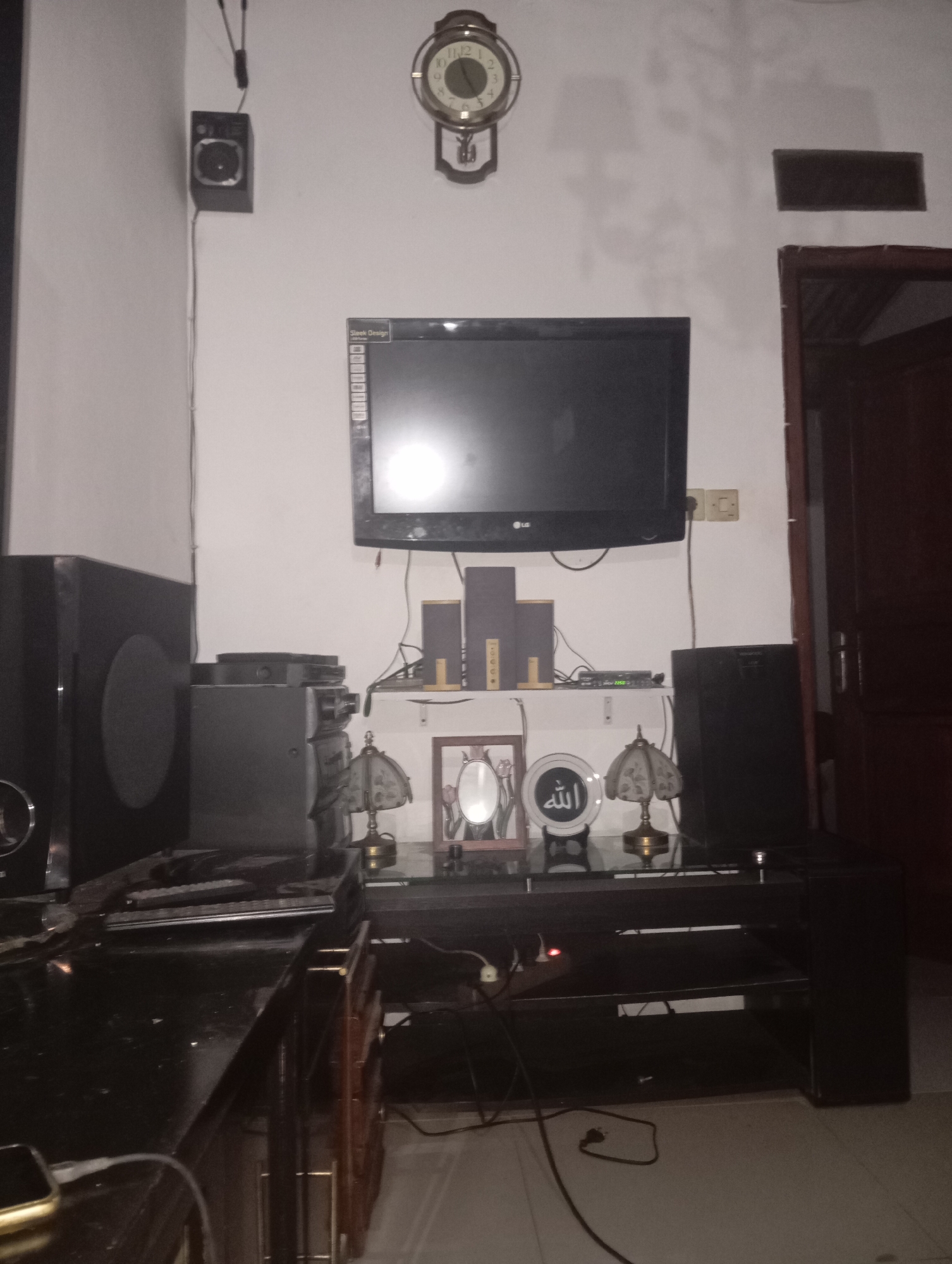 This is a forensic, matter-of-fact description of an indoor scene. The image shows a living room area with a television mounted on the wall. Below the TV, a dark entertainment unit holds various electronic devices, including speakers, a DVD player, and two lamps. A decorative plate with Arabic calligraphy and a framed picture are also on the unit. To the left of the entertainment unit, a large black speaker is visible. On the wall above the television, a round clock with Roman numerals is mounted. A smaller speaker is mounted on the wall to the far left. The room appears to be dimly lit. The overall setting suggests a typical domestic environment in Kedungbenda, Indonesia.