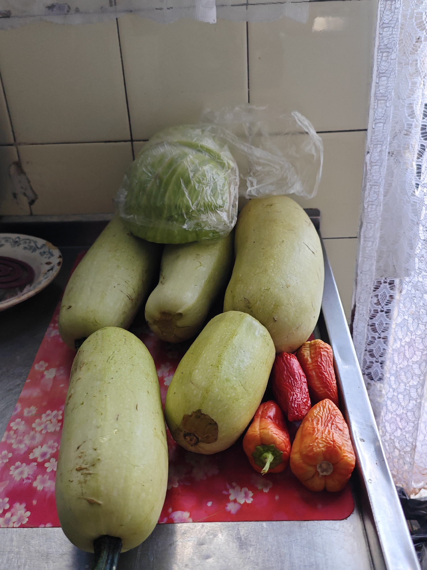 A close-up, high-angle, eye-level shot shows a collection of fresh vegetables on a metal surface, likely a kitchen counter, in Yartsevo, Russia. In the foreground, a large, light-green squash lies horizontally on a red cutting mat adorned with a floral pattern. To its right, another similar squash is positioned vertically, showing a brown blemish. Behind these, two more long, pale green squashes are clustered together. To the right of these squashes, a group of small, wrinkled peppers in vibrant shades of red and orange are arranged. In the background, a head of green cabbage is partially visible, wrapped in clear plastic. The wall behind the vegetables is tiled with pale yellow squares, and a white lace curtain hangs to the right. The overall impression is a still life of produce ready for preparation.