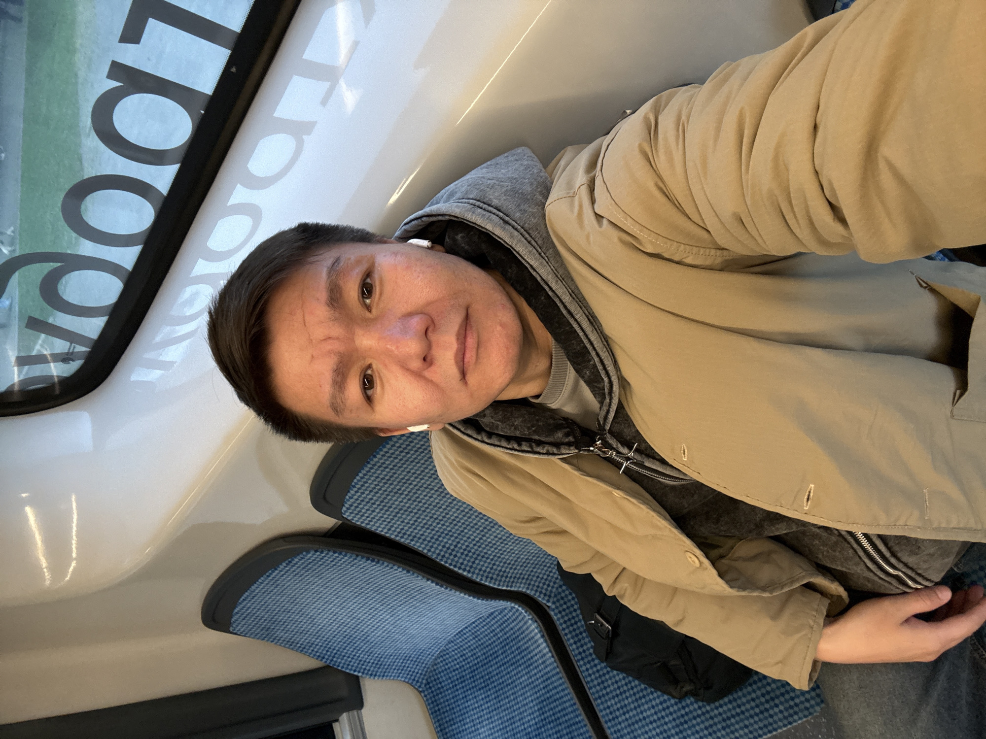 A man is seated on public transport, presumably a train, as the word 'Look' is visible in reverse through the window. The man is wearing a beige jacket over a grey hoodie and appears to be wearing earbuds. The seating is blue and patterned. The location is likely Moscow, Russia.