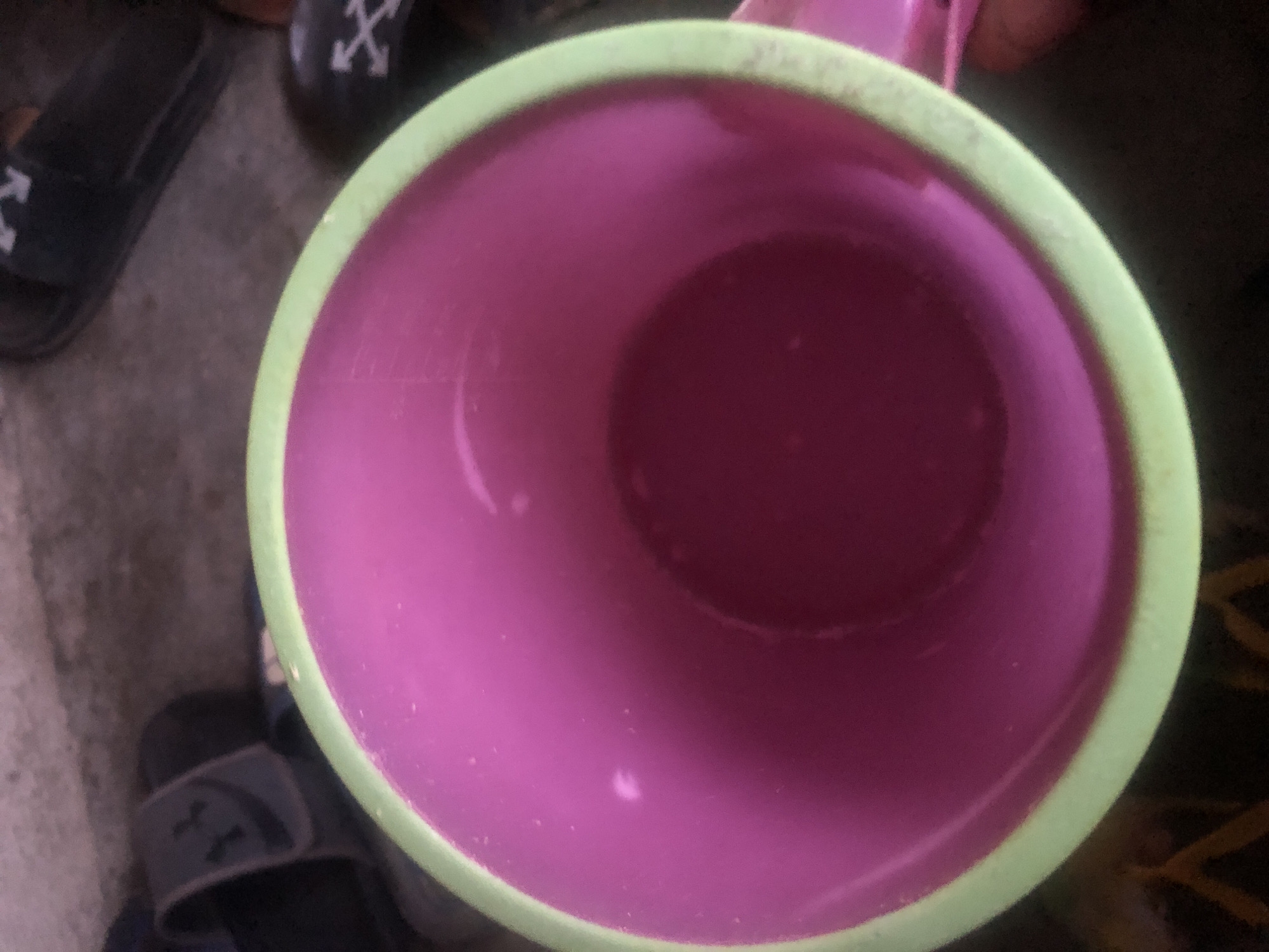 A close-up, overhead view shows the inside of a pink mug with a lime green rim, resting on a dark, textured surface. The mug appears to be made of plastic or a similar material. The interior of the mug contains a small amount of liquid, which is darker than the mug's interior. There are a few small white specks visible on the interior walls of the mug, possibly residue. In the background, out of focus, are dark colored sandals with white arrow logos, suggesting an outdoor or casual setting. The lighting is dim, contributing to the overall dark tones of the image, except for the bright pink and green of the mug. The scene is not specific to Jalingo, Nigeria; it's a generic depiction of a mug in an environment where sandals are present.