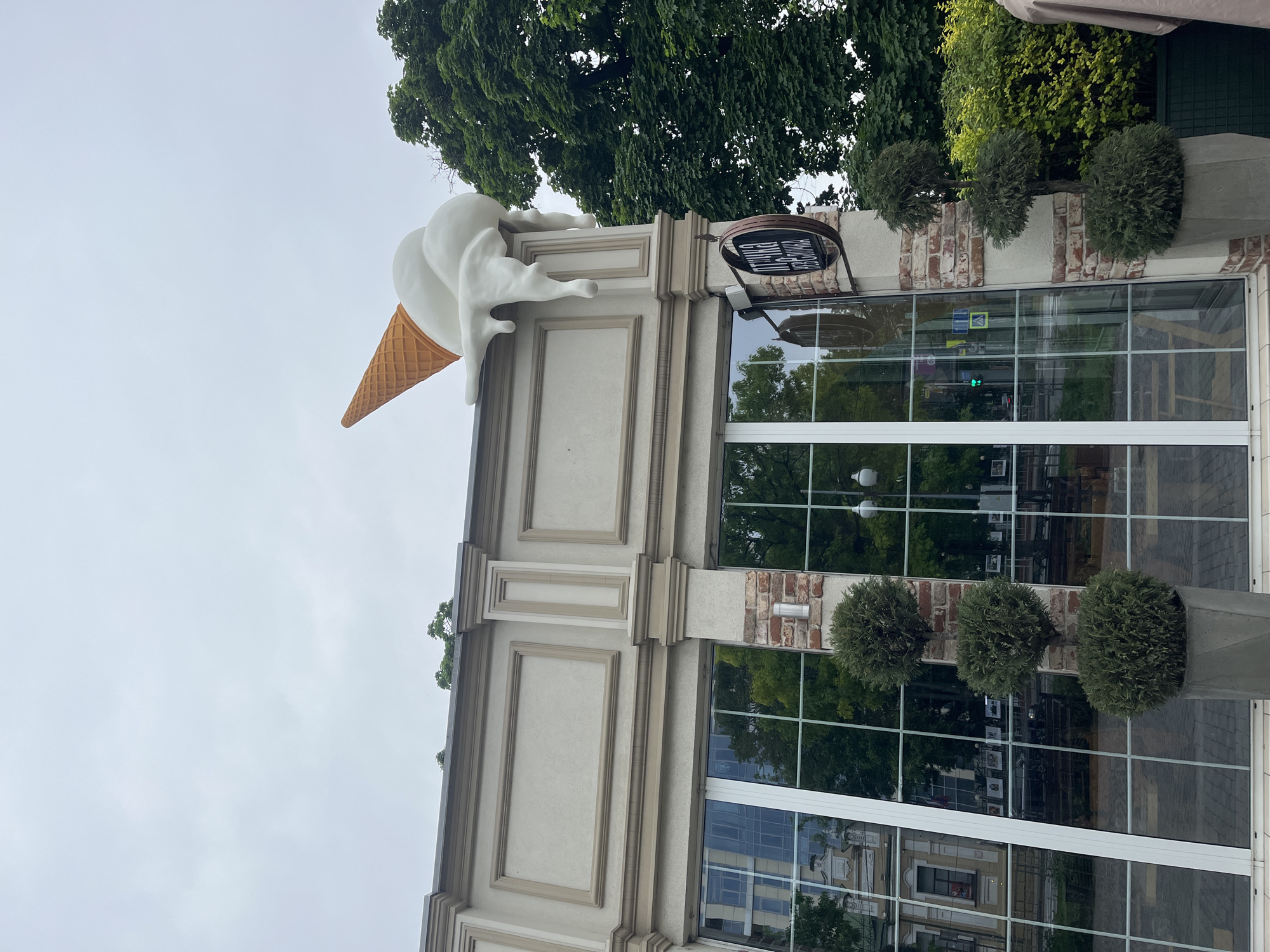 A low-angle view of a building, probably a shop, with a giant ice cream cone on the roof. The building facade is a mix of white, light tan, and brick. It has large paneled windows and round shrubs in planters. The sky is overcast. Reflections in the windows show street details and suggest the location is Krasnodar, Russia.