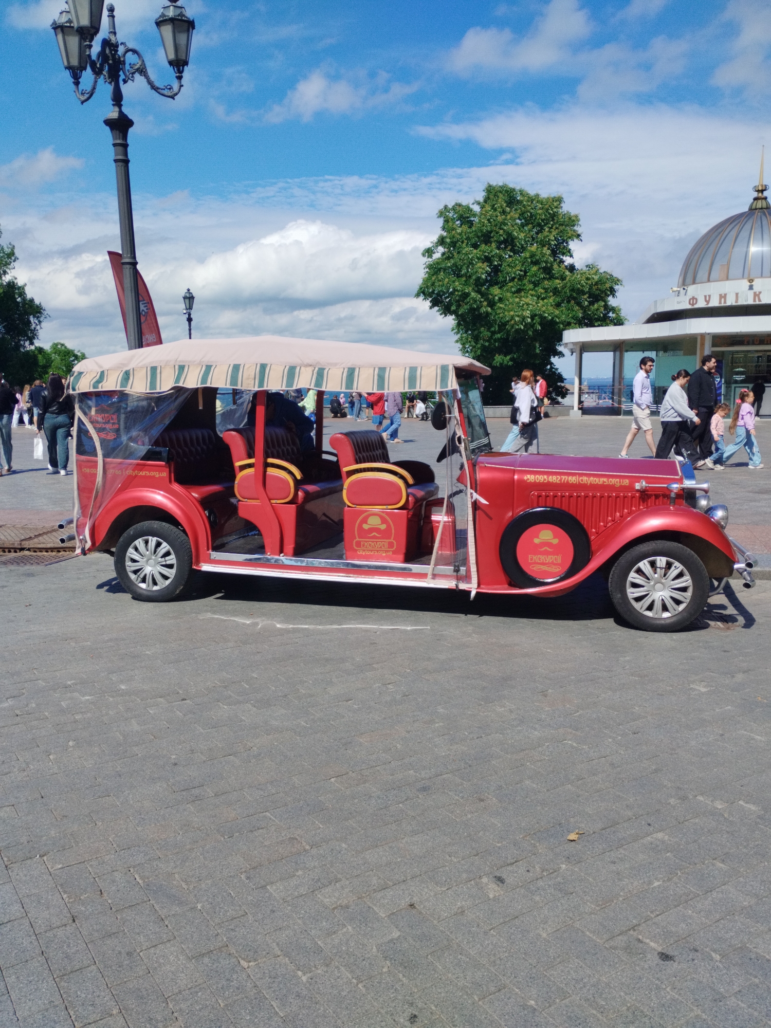 The image shows a red, antique-style tourist vehicle parked on a gray brick-paved surface. The vehicle has a striped beige and green canopy and red seats. A sticker on the vehicle displays the words "+38 093 482 77 66 citytours.org.ua" and "EKCKYPCII citytours.org.ua".
In the background, people are walking on a paved area, and behind them is a building with a domed roof labeled "ФУНІК" (Funicular). A tree is visible to the left of the building. A vintage lamppost stands to the left of the vehicle, and a red banner with white text is attached to it. The sky is blue with scattered white clouds.
This scene is located in Odesa, Ukraine, at the funicular station.