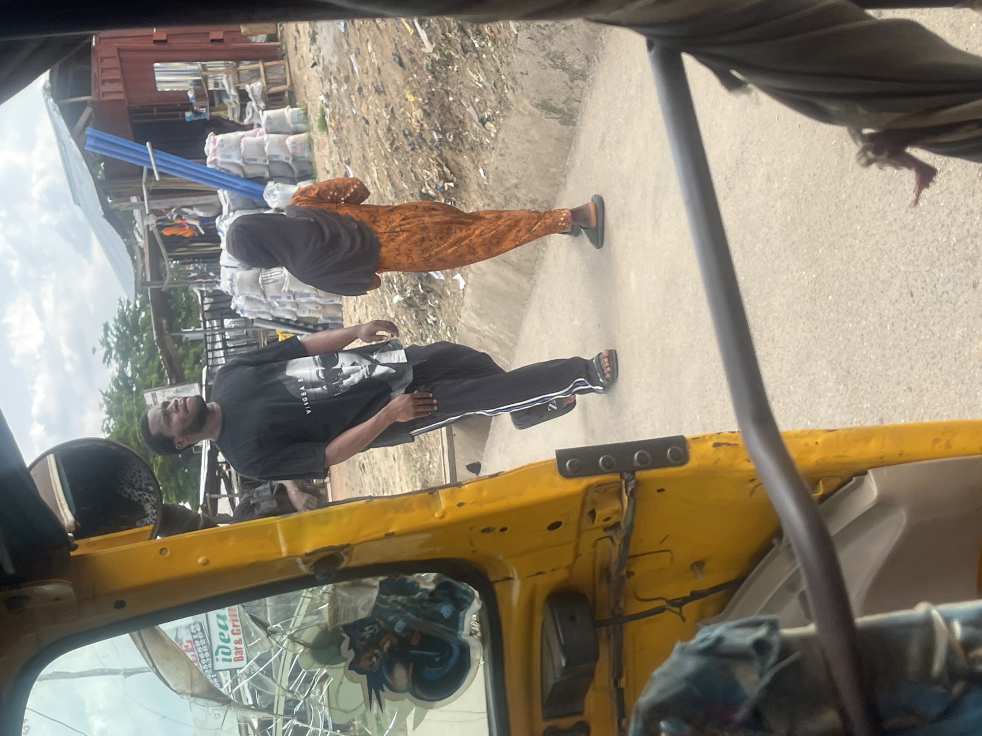 The image captures a street scene in Abuja, Nigeria, viewed from within a yellow vehicle, likely a tuk-tuk. In the foreground, the interior of the vehicle is visible, with a dark-colored metal bar extending across the lower portion of the frame. The yellow exterior of the vehicle is also prominent on the left side, featuring a cracked window with stickers on it, including one depicting a motorcycle.
Two individuals are the main subjects in the middle ground. A Black man wearing a black t-shirt with a graphic print and black pants with white stripes is walking away from the camera. His t-shirt displays a black and white portrait. He is wearing sandals. Beside him, slightly ahead and to his right, a person wearing an orange jumpsuit and a dark headscarf is also walking away. This individual is wearing dark-colored slides.
The environment appears to be a dusty, unpaved area bordering a street. In the background, there are several structures that resemble market stalls or makeshift shops. One building has a corrugated metal roof and is constructed with a red shipping container and wood. Piles of materials, including what appear to be sacks of cement or similar products and blue pipes, are stacked near the structures. Debris and trash are scattered on the ground. The sky above is overcast. The overall impression is of a busy, informal commercial area.