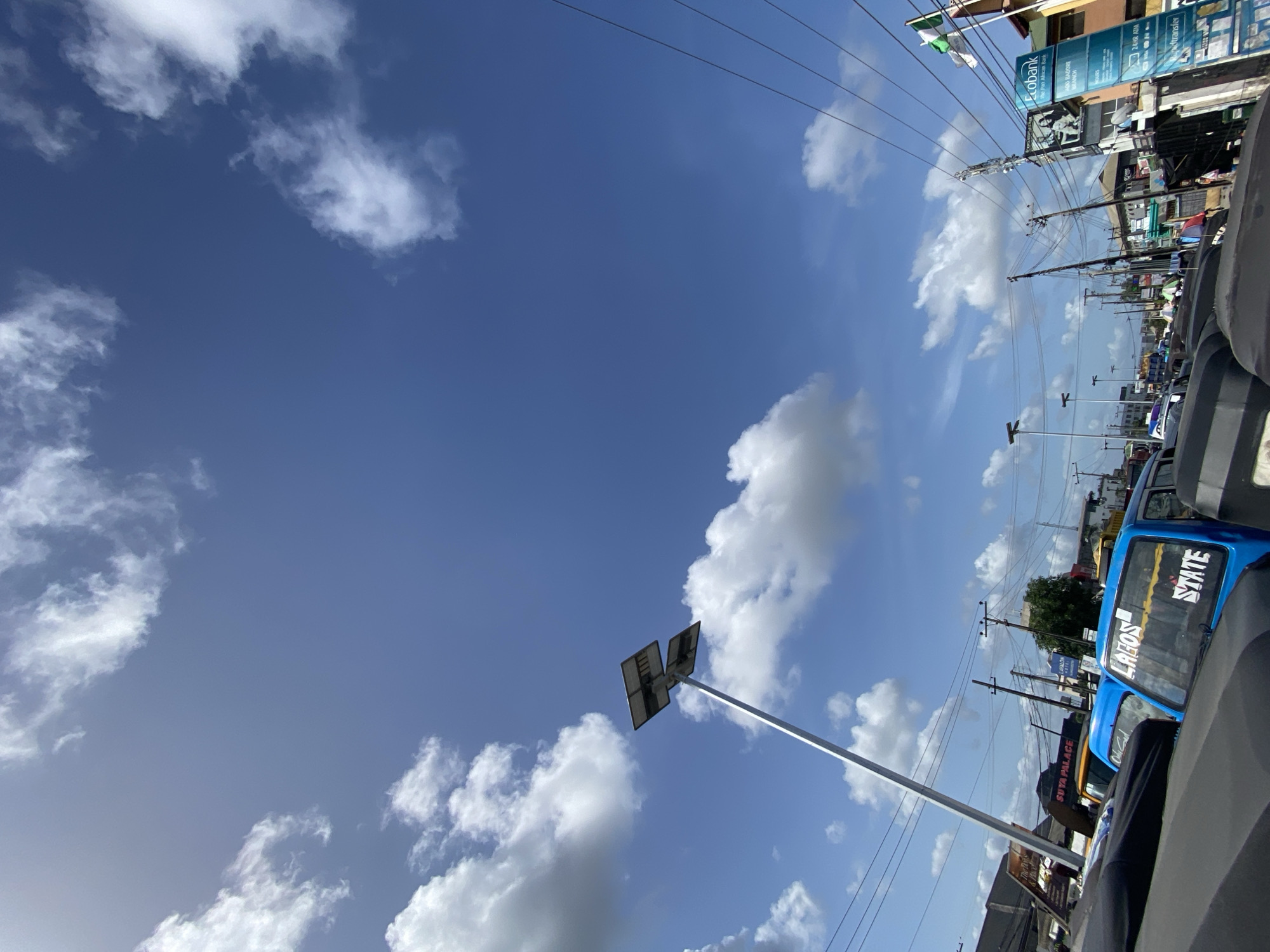 The image shows a view of Ado, Nigeria, under a bright blue sky with scattered cumulus clouds. Utility poles and power lines run across the sky. Several vehicles, including commercial tricycles ("keke"), are visible in the foreground. Signs for Ecobank, SUYA PALACE, LAVALON, and a "LAGOS STATE" labeled tricycle are visible.