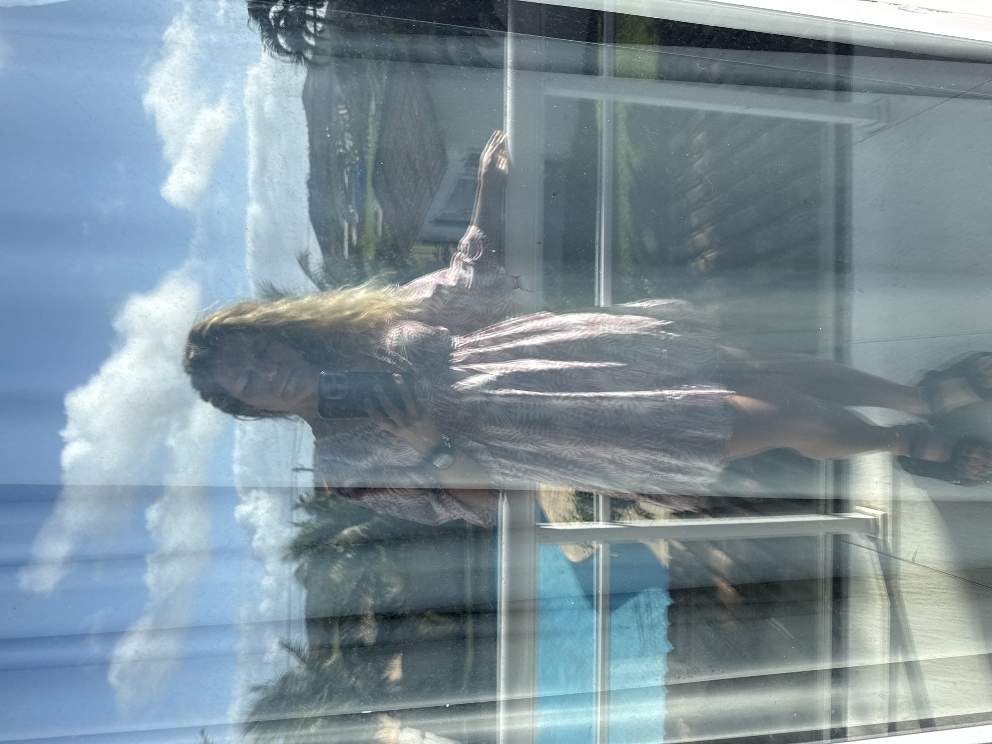 A woman with blonde hair, wearing a short pink and white patterned dress and dark sandals, stands on a balcony, taking a mirror selfie with a smartphone. She is holding the phone in her right hand, and her left hand rests on a white railing. The balcony overlooks a swimming pool and a landscape with palm trees and hills. The scene appears to be in Cabo Frio, Brazil, suggested by the tropical vegetation and architecture.