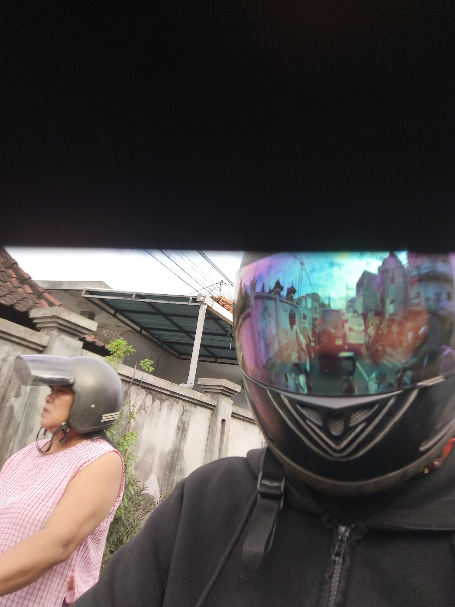 Two individuals are captured in a photograph, seemingly on a motorcycle. The person in the foreground is wearing a black hooded jacket and a black helmet with a reflective visor. The visor reflects a busy street scene with vehicles and buildings. To the left, another person, a woman, is visible from the chest up. She is wearing a light pink and white checkered sleeveless top and a dark gray helmet with a clear visor. The background shows a concrete wall and a building with a tiled roof. Power lines are visible overhead. The lighting suggests it is daytime.