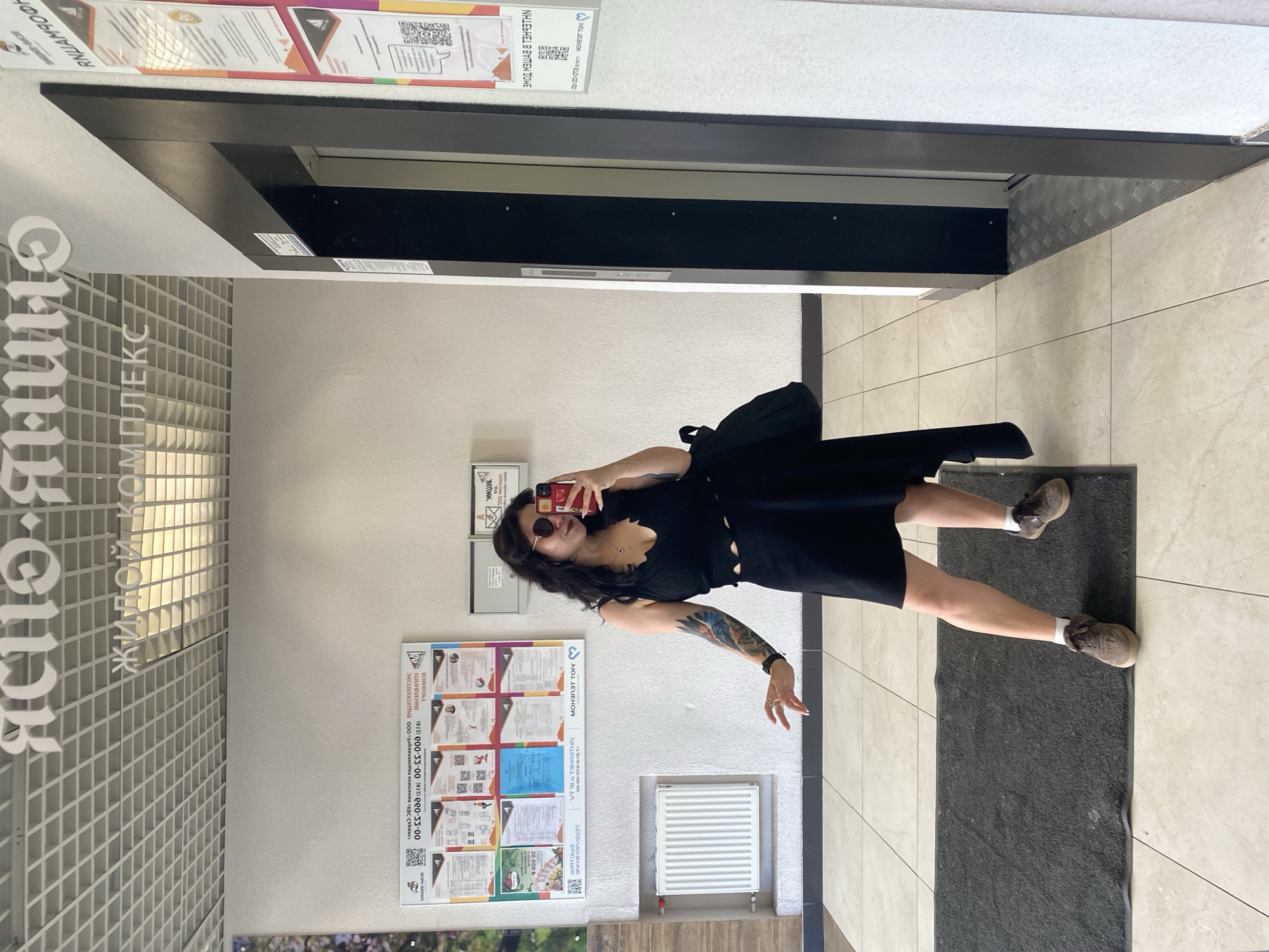 A woman is taking a mirror selfie in the lobby of a residential complex in Yanino-1, Russia. She is wearing a black sleeveless top and a black skirt, paired with white socks and brown sneakers. She has sunglasses on and is holding her phone in her right hand. A black bag is slung over her left shoulder. Her left arm, which is visible, has a tattoo. The background shows a tiled floor, a radiator on the wall, a bulletin board with various documents and flyers, and an elevator. Above the elevator, the text "ЯСНО.ЯНИНО" and "ЖИЛОЙ КОМПЛЕКС" are visible. The overall environment appears to be a modern residential building lobby.