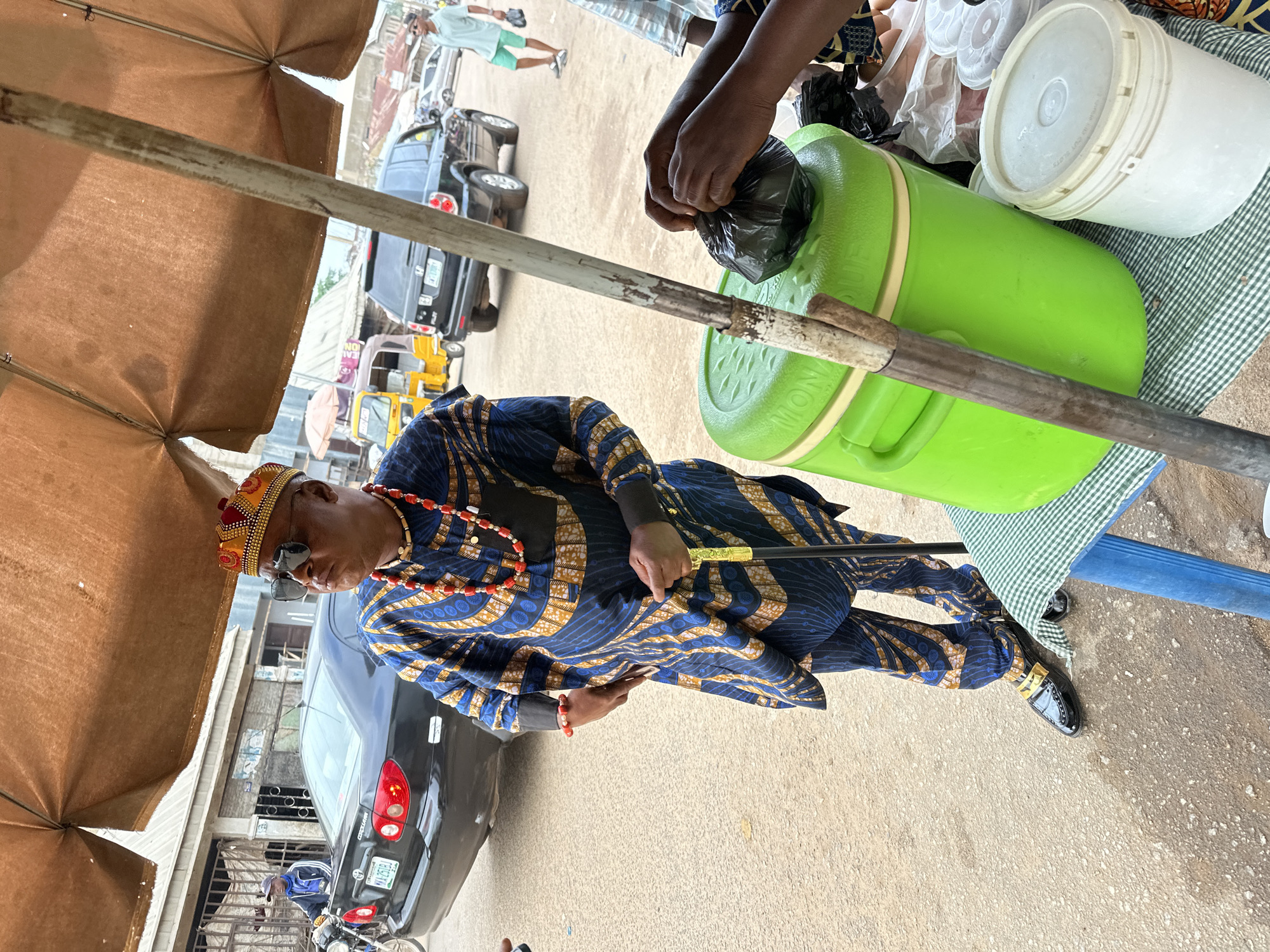 In the city of Narayi, Nigeria, a man stands under a brown canopy, dressed in a traditional blue and gold patterned outfit. He wears a decorative red and gold beaded crown, sunglasses, and a multi-strand red beaded necklace. He holds a gold-topped cane in his right hand. In front of him, on a table covered with a checkered cloth, are two large containers: a bright green one and a white one. A person's hands are visible, tying a black plastic bag. In the background, a black car is parked on a dusty street, with other vehicles and buildings suggesting an urban environment.