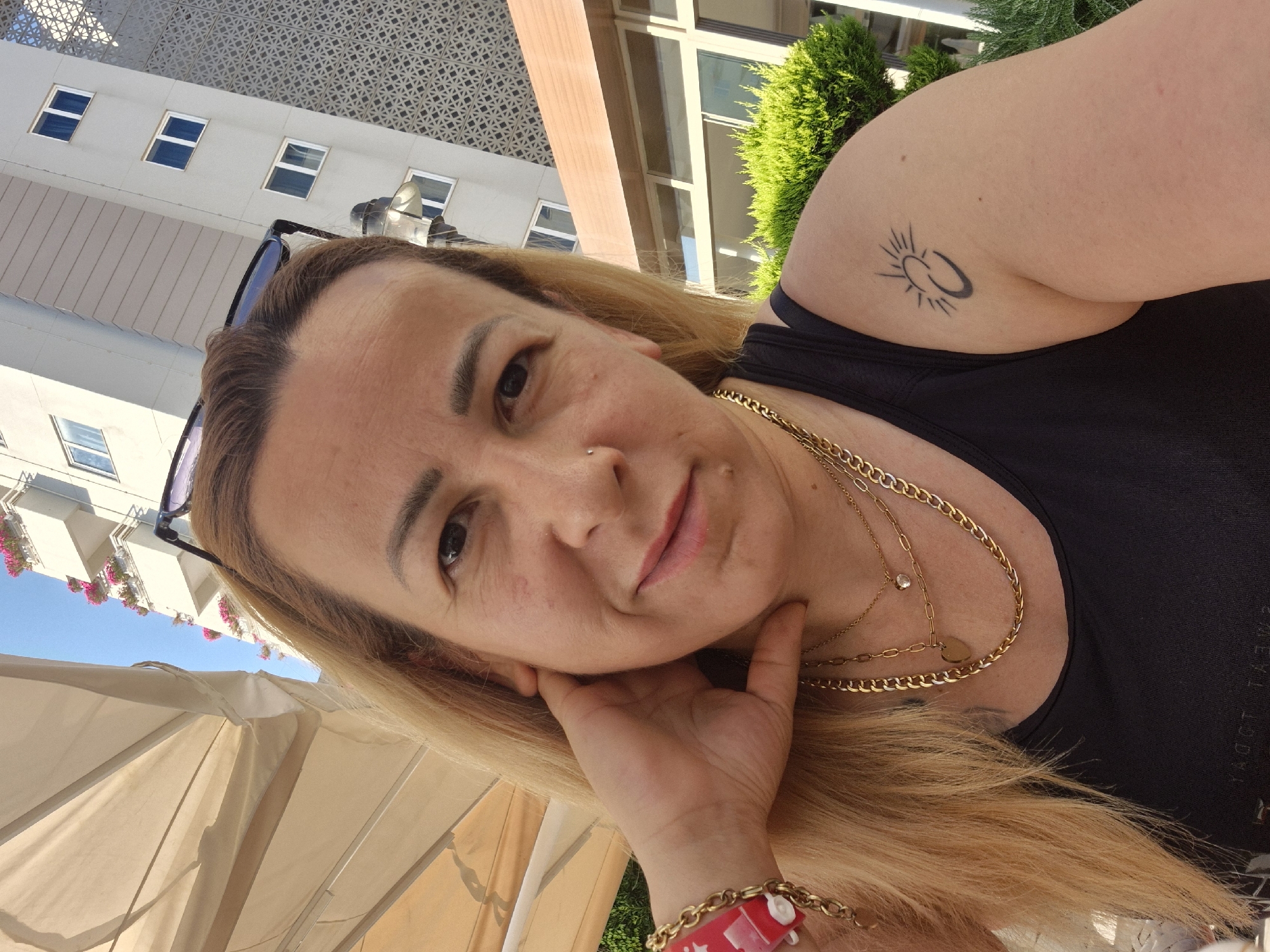 In an outdoor, urban setting, a woman with blonde hair and light skin takes a selfie. She is wearing a black tank top, a gold chain necklace with a heart pendant, and a red bracelet. Her left hand is resting on her face. On her right arm, there is a tattoo of a sun with a crescent moon. In the background, a modern building with multiple windows and decorative panels stands under a clear blue sky. There is also a large beige awning and green bushes. The scene suggests a sunny day, possibly in a tourist area or resort.