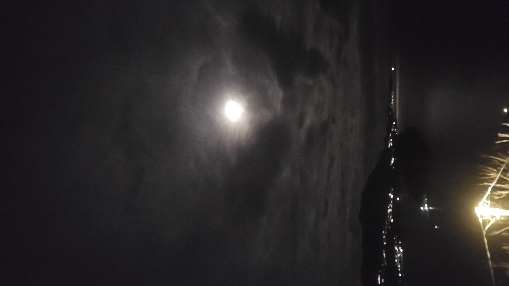The image shows a nighttime scene with a bright moon partially obscured by clouds, providing illumination to a body of water. A dark, vertical object is positioned on the right, possibly a structure or part of a building, with reflective points suggesting lights or water droplets. There are faint traces of what appear to be fireworks in the far right corner. Given the details, the location is most likely in or around Budva, Montenegro, with the body of water being the Adriatic Sea. The vertical object is probably a building or structure situated along the coast.