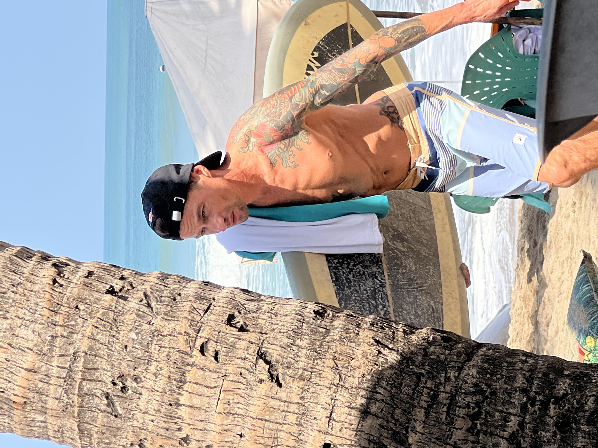 A shirtless man with extensive tattoos on his arm and torso walks on a sandy beach in Kuta, Indonesia, carrying a surfboard under his right arm. He is wearing a black baseball cap and striped swim trunks. A white towel with a teal stripe is draped over his left shoulder. In the background, the turquoise ocean meets the sky, with a white umbrella and green plastic chairs visible on the beach. A palm tree trunk is in the foreground on the left side of the image. The scene appears to be a sunny day.