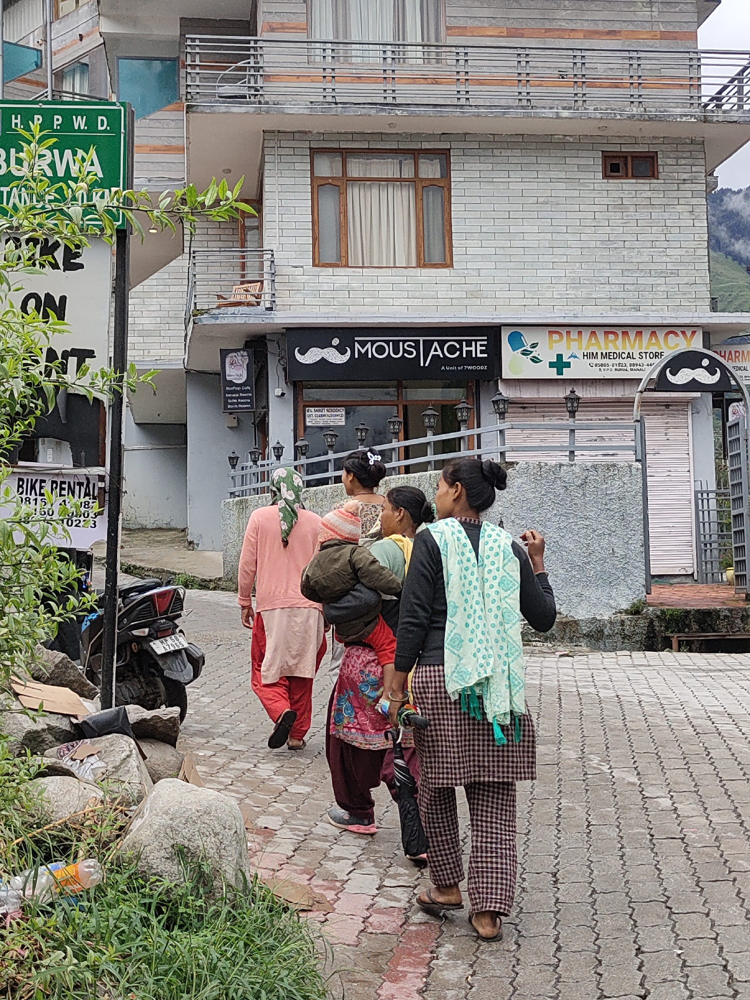 The image depicts a street scene in Burwa, India, with a building in the background featuring multiple stories and balconies. A green sign with white text "H. P. P. W. D." and "BURWA" is visible on the left side of the frame. Below this, a white sign with black text indicates "BIKE RENTAL" and provides a phone number.
In front of the building, three individuals are walking away from the camera on a paved walkway. The person closest to the camera is wearing a checkered outfit and a light blue scarf with a patterned design. The second person is wearing a dark jacket and a pink headscarf, carrying a bundle on their back. The third person is wearing a pink top and red pants. A child in a red and pink outfit is partially visible behind the second person.
Several shops are located on the ground floor of the building, with signs that read "MOUSTACHE" and "PHARMACY HIM MEDICAL STORE". A motorcycle is parked on the left side of the walkway. The ground is wet and appears to be paved with interlocking bricks. Rocks and green foliage are present on the left side of the image. The overall environment suggests an urban or semi-urban setting in a hilly region, possibly during or after rainfall.