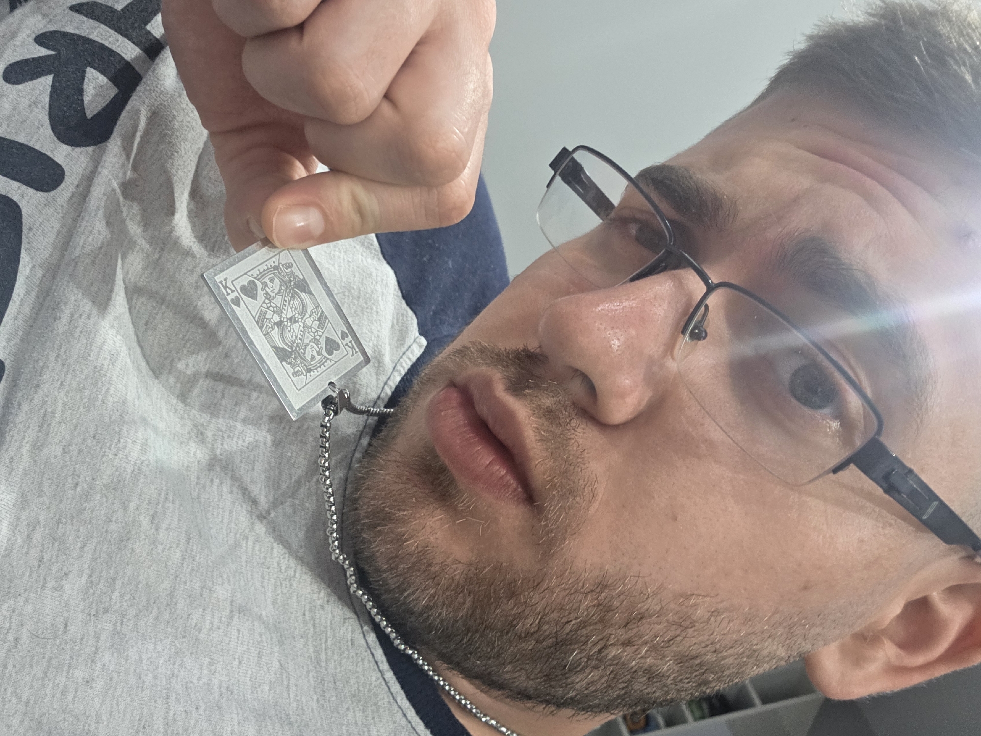 A close-up, angled shot shows a man holding a playing card charm (King of Hearts) suspended from a chain. The man has short brown hair, a beard, and is wearing glasses. He is wearing a grey and blue hooded sweatshirt. The background is slightly blurred, but what appears to be shelving is partially visible. The location in the city of Yagodovo, Bulgaria is undetermined.