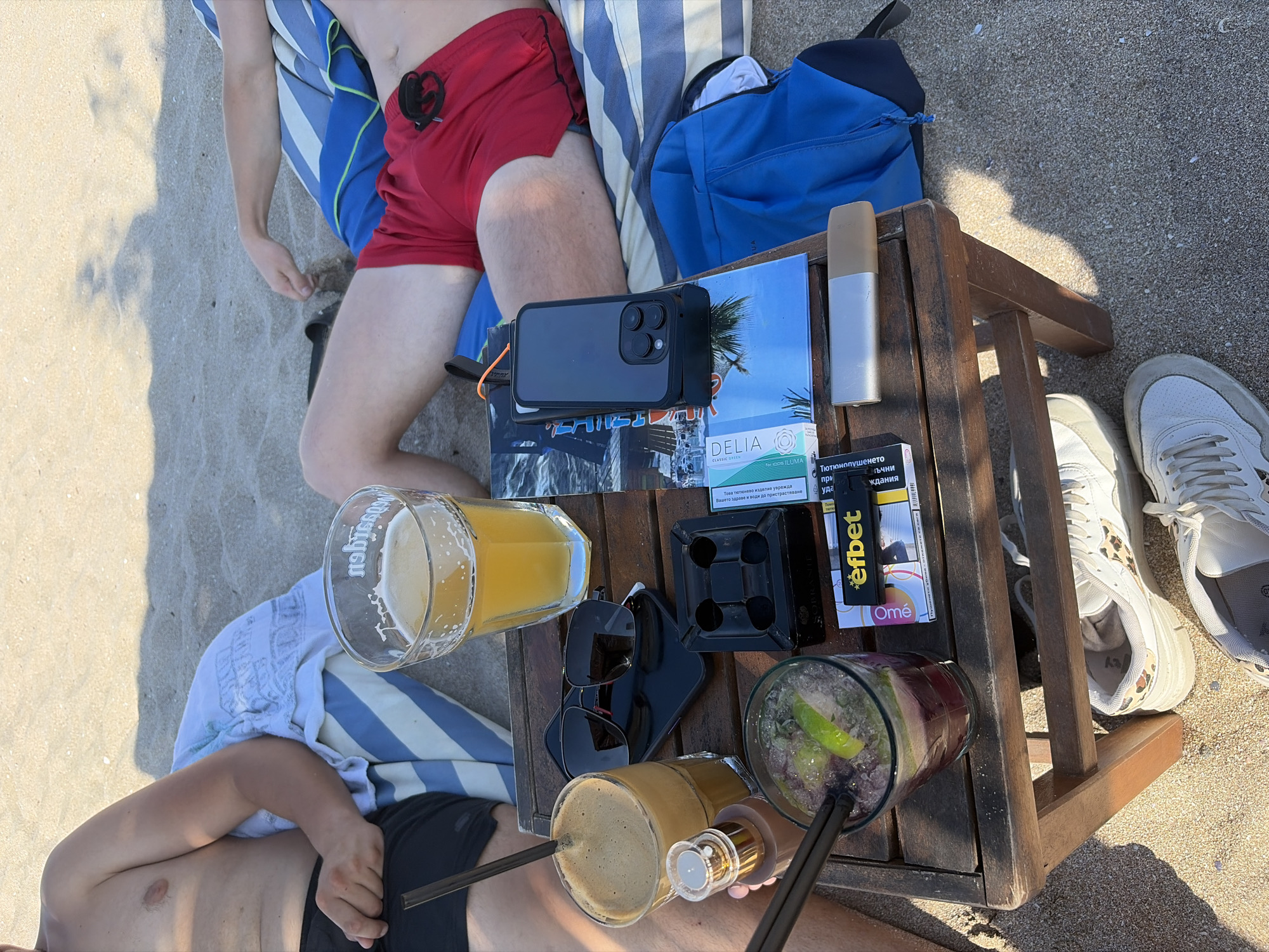 The image depicts a beach scene in Lozenets, Bulgaria, on a sunny day. Two men are relaxing on the sand, seated on striped towels. In the foreground, a wooden table holds several items, including two drinks, one with ice and lime, another frothy beverage, a glass of beer, sunglasses, an ashtray, a mobile phone, a package of cigarettes, and an electronic smoking device. The men appear to be enjoying leisure time, possibly at a beach bar or cafe. The environment is characterized by sand and the presence of beach towels, suggesting a typical seaside setting. The people are dressed casually, in swim shorts and what appears to be a bare chest. The overall impression is one of relaxation and summer enjoyment.