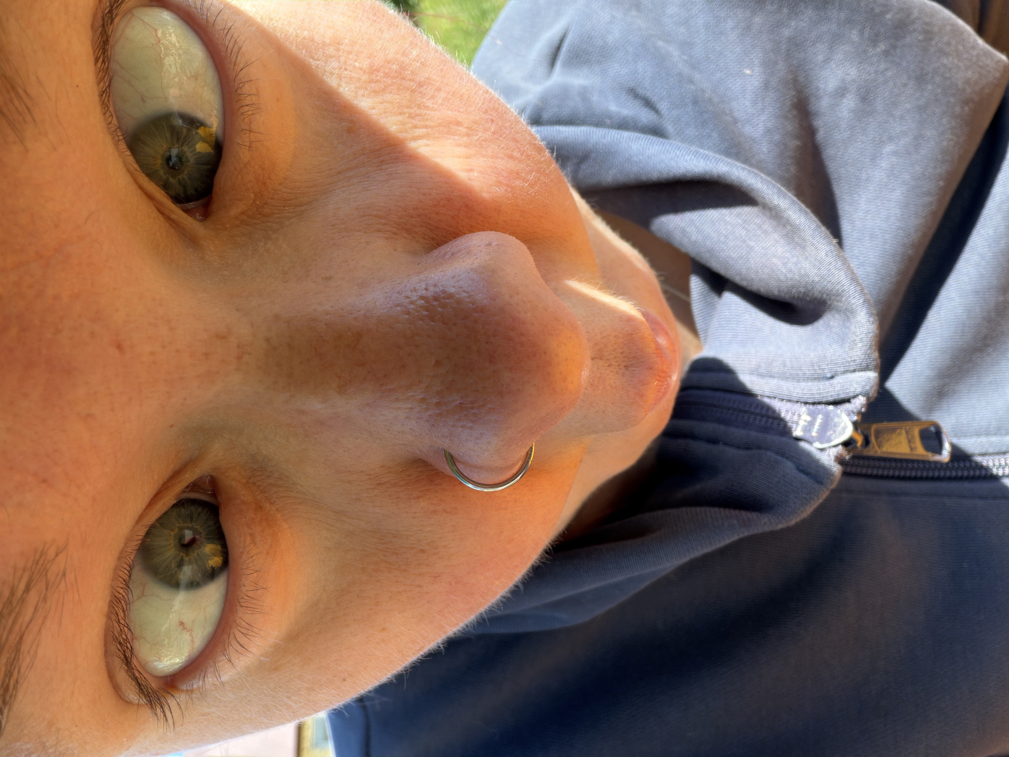 The image is a close-up, low-angle shot of a person's face, focusing on their eyes and nose. The individual has light skin with some visible pores and a few freckles on their nose. Their eyes are wide open, with green irises and dark pupils. There appear to be some small, yellowish specks within the irises. The person has a silver hoop piercing in their right nostril. They are wearing a dark blue hooded sweatshirt with a zipper that is partially visible. The lighting suggests an outdoor environment, with sunlight casting shadows and highlighting certain features of the face. The background is blurred, but appears to be green, possibly foliage or grass. There is no specific indication of a location within the city of Chaikovsky, Russia, or any other geographical context.