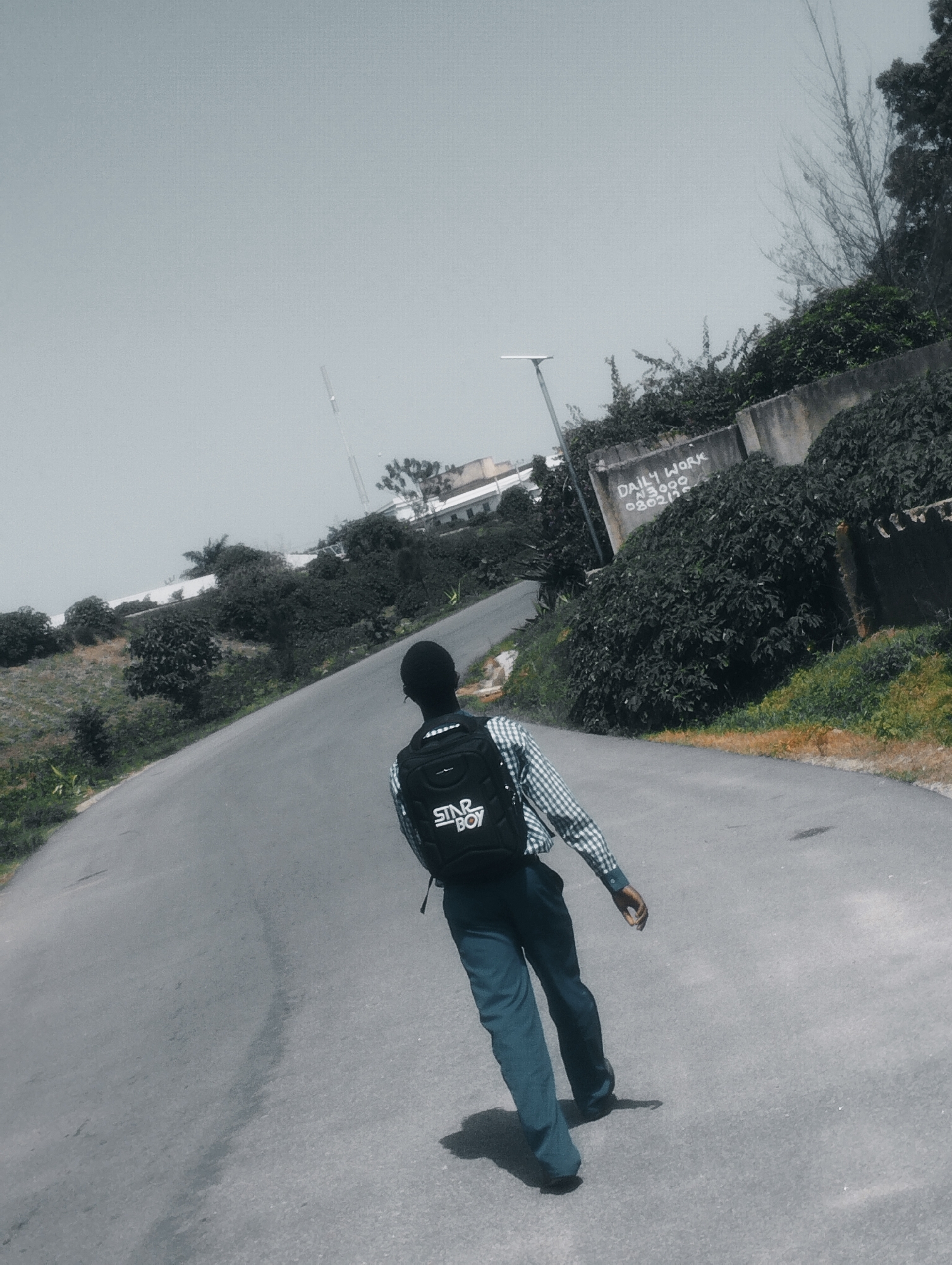 A person walks away from the camera on an asphalt road. They are wearing a checkered shirt and dark pants, with a black backpack on their back. The backpack has the words "STAR BOY" printed on it. The scene appears to be outdoors, with vegetation on either side of the road. In the background, there are some buildings and a concrete wall with graffiti that reads "DAILY WORK N3,000 08030215". The sky is overcast, suggesting a cloudy day. The location is identified as Jos, Nigeria. The person's posture and the direction they are walking suggest they are on a journey or heading somewhere. The overall mood is neutral.