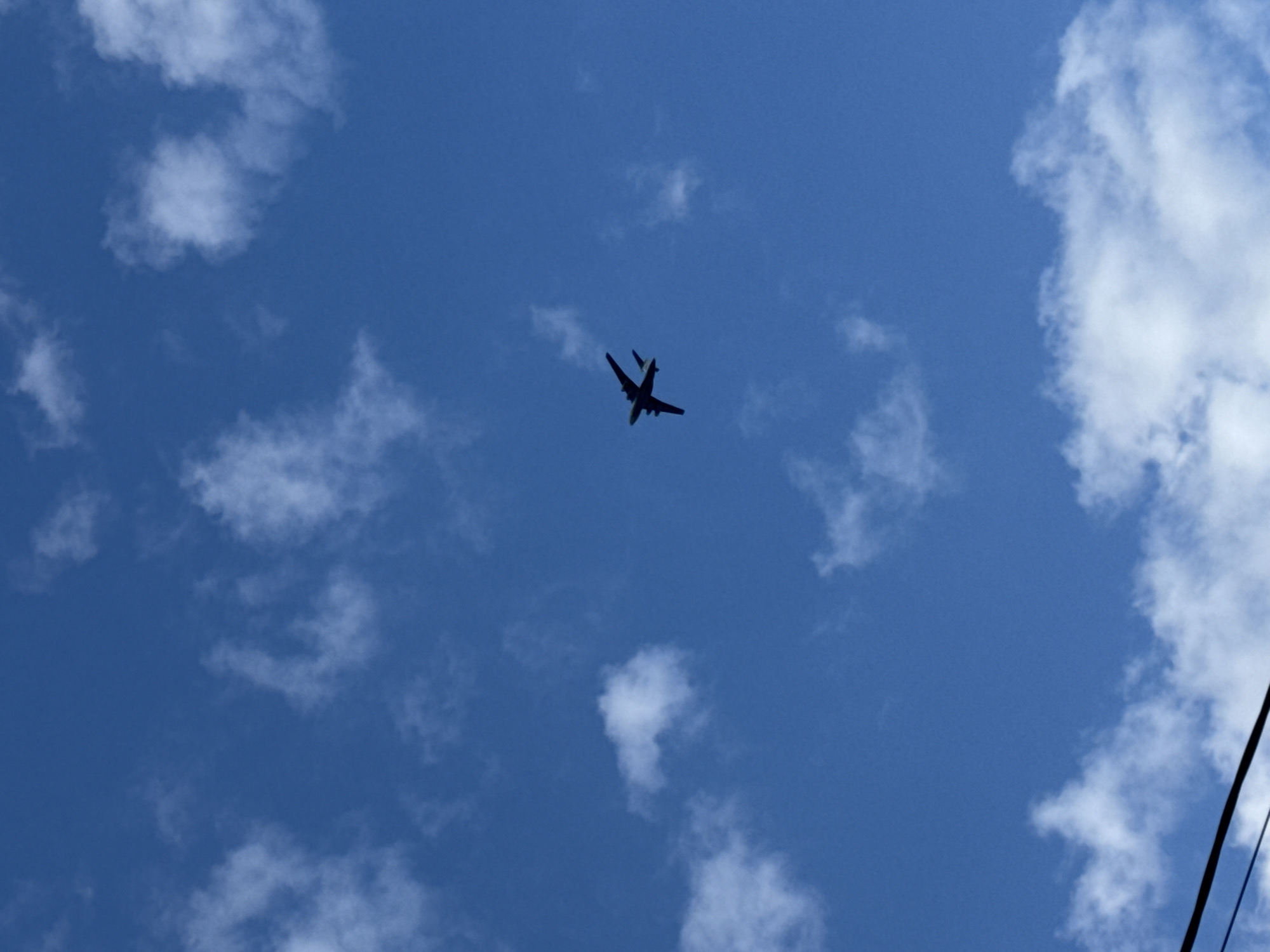 An upward-facing shot captures the silhouette of a jet aircraft soaring across a vibrant blue sky, dotted with scattered white clouds. The aircraft, with its swept wings and what appears to be a twin-tail configuration, is positioned towards the center of the frame, indicating it's flying overhead. Its dark outline suggests the sun is either behind or directly above the plane, making identification of specific features difficult.
The sky is largely clear, with several patches of fluffy, white cumulus clouds distributed throughout the upper and lower sections of the image. A distinct dark, diagonal line, resembling a power line or cable, is visible in the bottom right corner, suggesting the photo was taken from a ground-level perspective looking up. The overall impression is of a bright, clear daytime sky. The scene is set in Rostov-on-Don, Russia. No visible text is present in the image.
