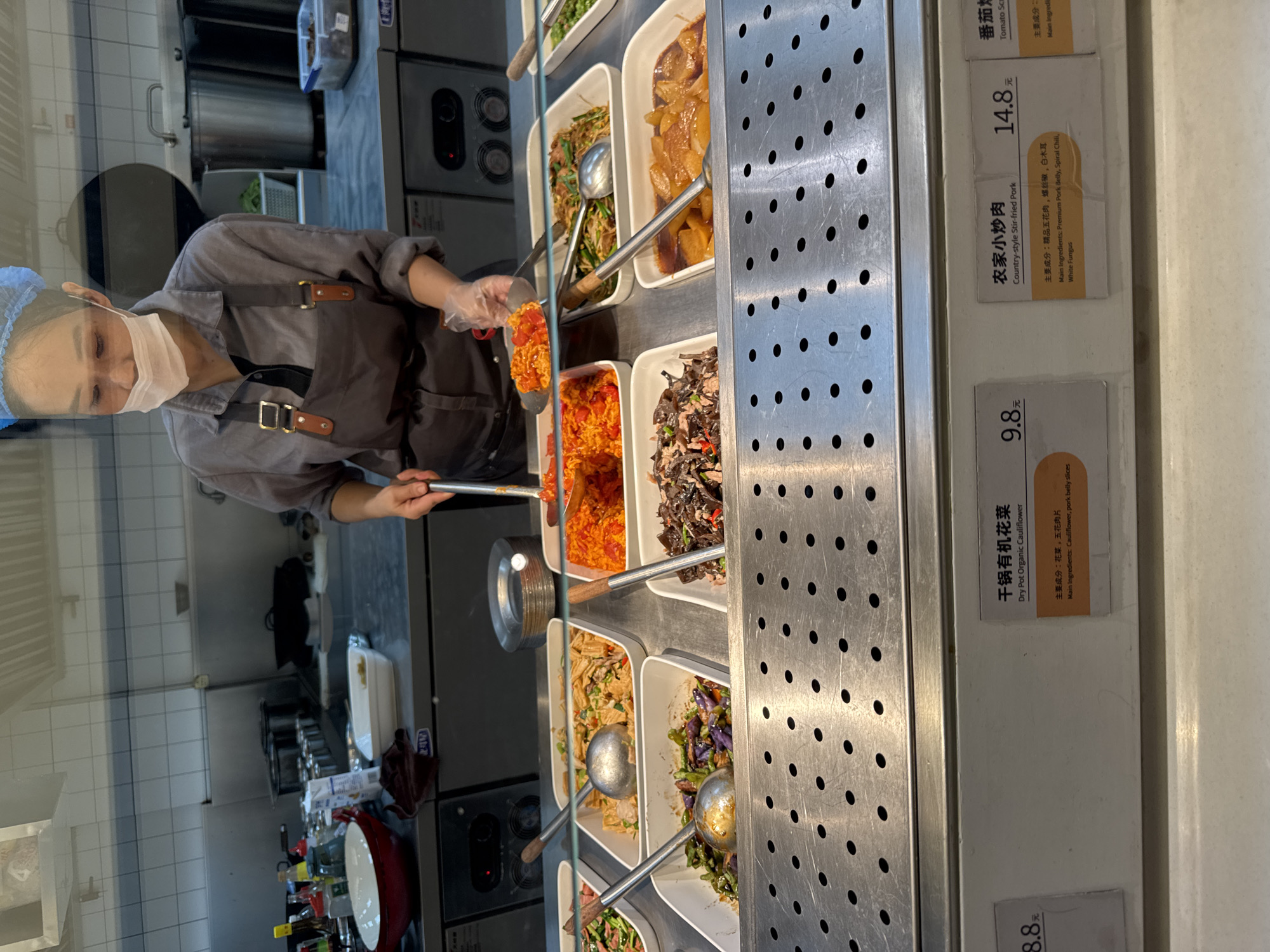 A woman wearing a hairnet, face mask, and apron is serving food at a buffet counter in a canteen. She is holding a serving spoon and a small metal plate filled with orange-colored food, possibly stir-fried tomatoes or peppers. The buffet counter displays various dishes in white rectangular containers, including what appears to be stir-fried vegetables, noodles, and meat. Signs with Chinese and English text are visible below some of the dishes, indicating their names and main ingredients. The environment appears to be a commercial kitchen or food service area, with stainless steel surfaces and kitchen equipment in the background. The location is identified as Yangpu District, China.