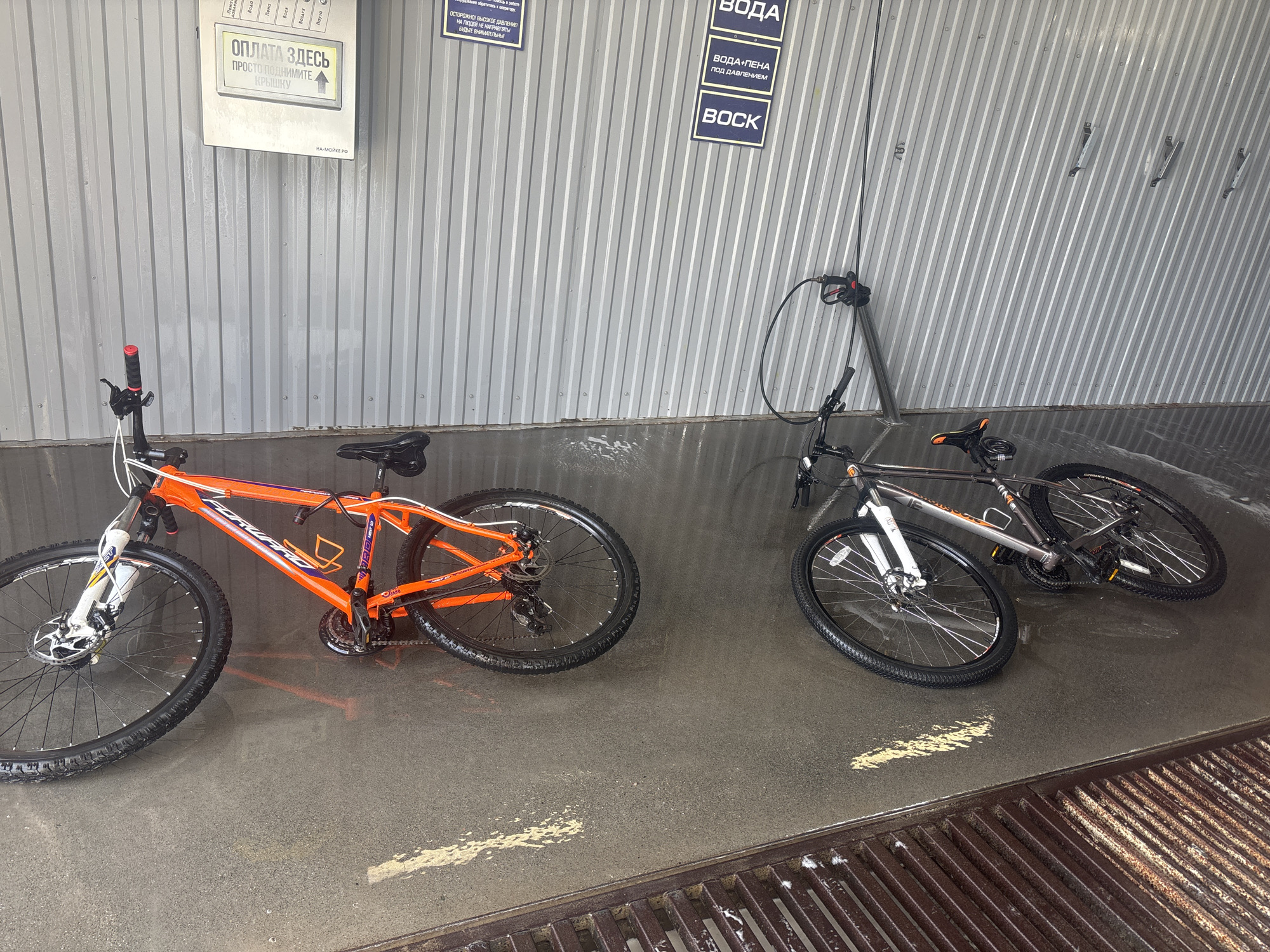 Two bicycles are parked on the wet, tiled floor of an outdoor car wash in Chaikovsky, Russia. The bicycle on the left is orange and white, with disk brakes. The bicycle on the right is silver and black, also with disk brakes. Above the bicycles, on a corrugated metal wall, are signs indicating different car wash services, including "WATER," "WATER-FOAM," and "WAX." A hose with a spray nozzle is visible hanging from a metal pole to the right of the bicycles. The floor is wet, with water puddles and foam visible. The overall scene suggests a self-service car wash facility.