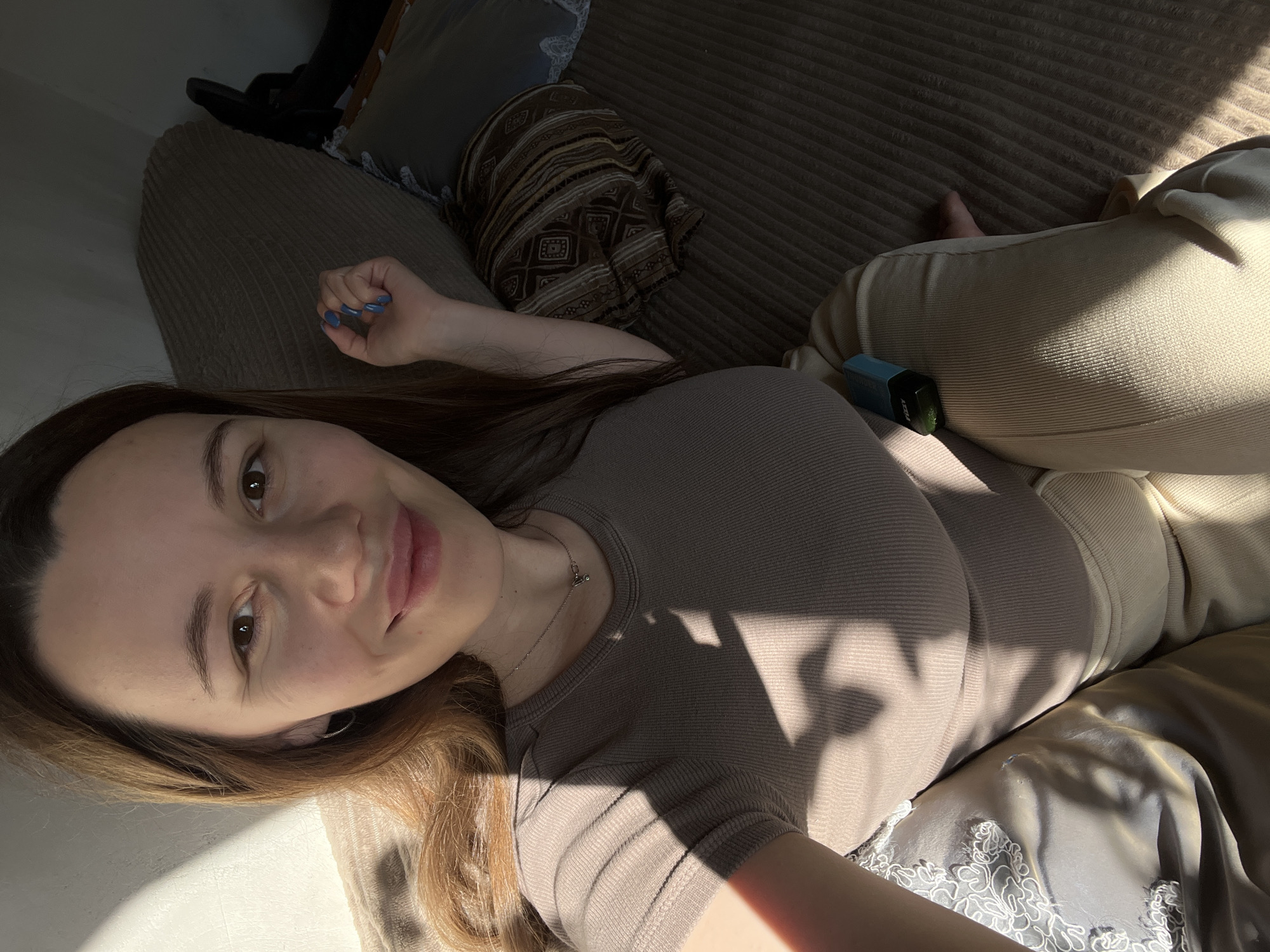 An indoor, overhead selfie depicts a female subject, appearing to be in her 20s or 30s, seated on upholstered furniture. She has long, straight brown hair, brown eyes, and a light complexion, displaying a subtle smile. Her left hand, with blue painted fingernails, rests on her upper thigh. She wears a form-fitting, ribbed, short-sleeve top in a light brown or beige color, a thin silver chain necklace with a small pendant, and a small silver hoop earring in her visible left ear. Her lower body is clad in light-colored, possibly beige corduroy trousers. A light blue and black electronic vaping device, branded "FIZZY," rests on her right thigh.
The subject is positioned on a ribbed, light brown/beige sofa or bed. To her immediate left is a cushion with grey fabric and white lace embroidery. Behind her to the right, a pillow with a dark brown and beige geometric pattern is visible, along with a plain dark grey pillow partially obscured. Strong directional sunlight enters from the left, casting distinct, sharp shadows across her face, torso, clothing, and the furniture, creating diagonal patterns. The background to the upper left features a plain, light-colored wall.