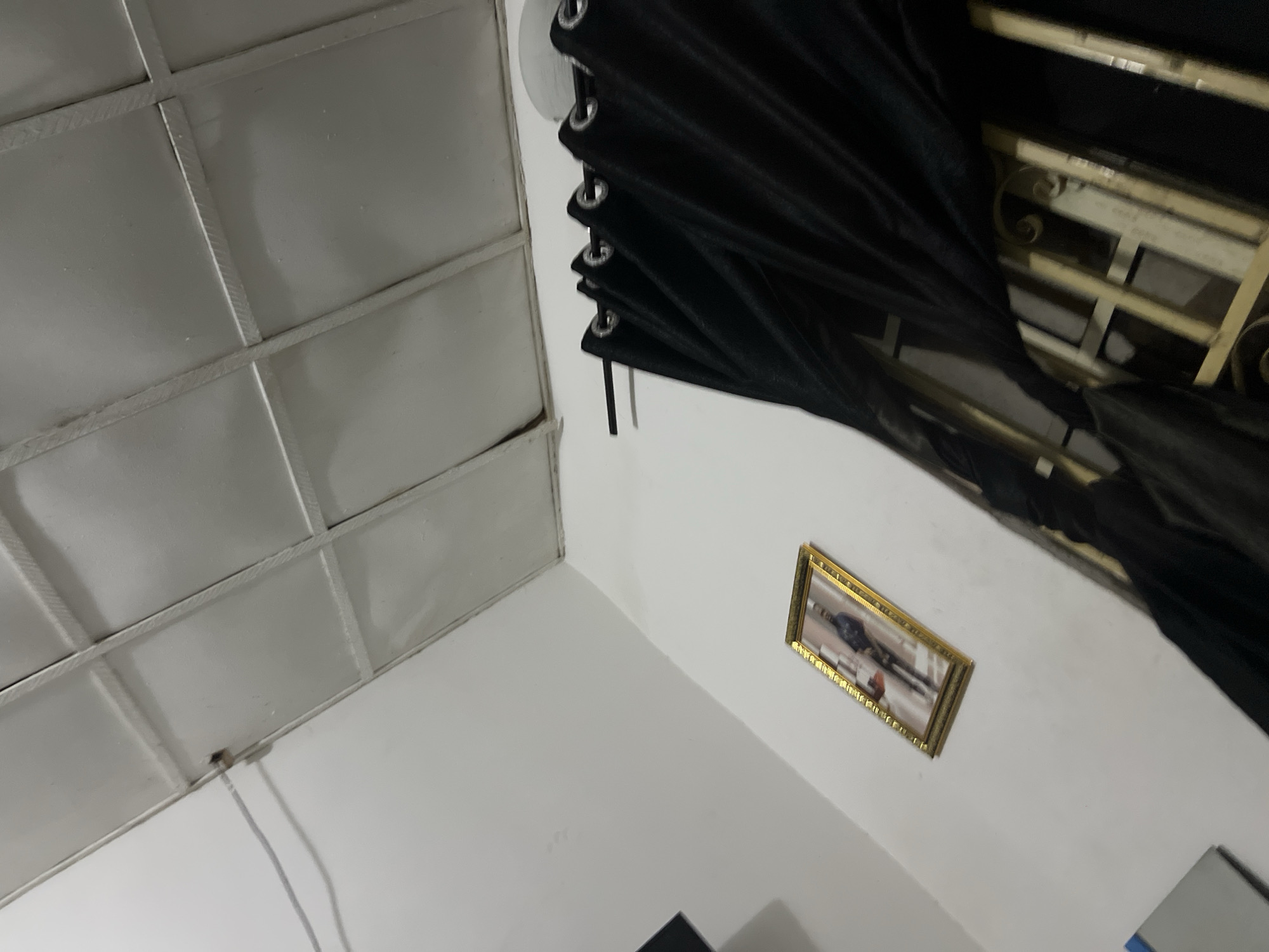 The image shows an interior view of a room, possibly in Howayi, Nigeria. The room features a white, grid-patterned ceiling with a visible wire running across it. On a white wall, a framed picture hangs, depicting a person in a dark jacket. To the right, black curtains with grommets are drawn across a window with metal bars. The overall lighting is dim.