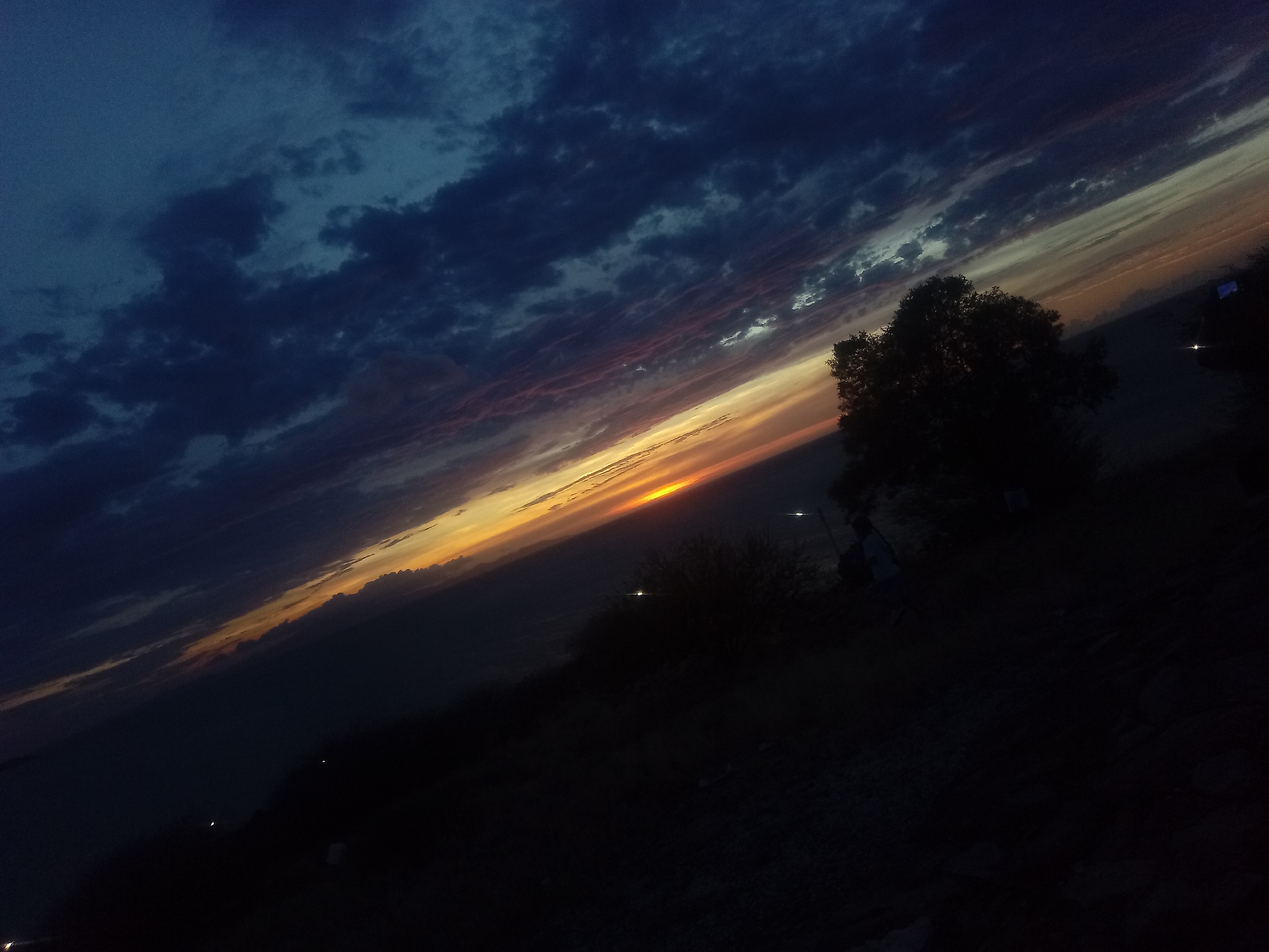 The image shows a dark, angled view of a sunset from what appears to be a hillside overlooking Juan Griego, Venezuela. The sky is dominated by dark blue and gray clouds in the upper left, transitioning to a vivid orange and yellow sunset near the horizon. A silhouette of a tree is visible on the right side of the frame. The foreground is mostly dark and indistinct, showing signs of vegetation and rocky terrain. Some distant lights suggest activity or structures near the horizon.