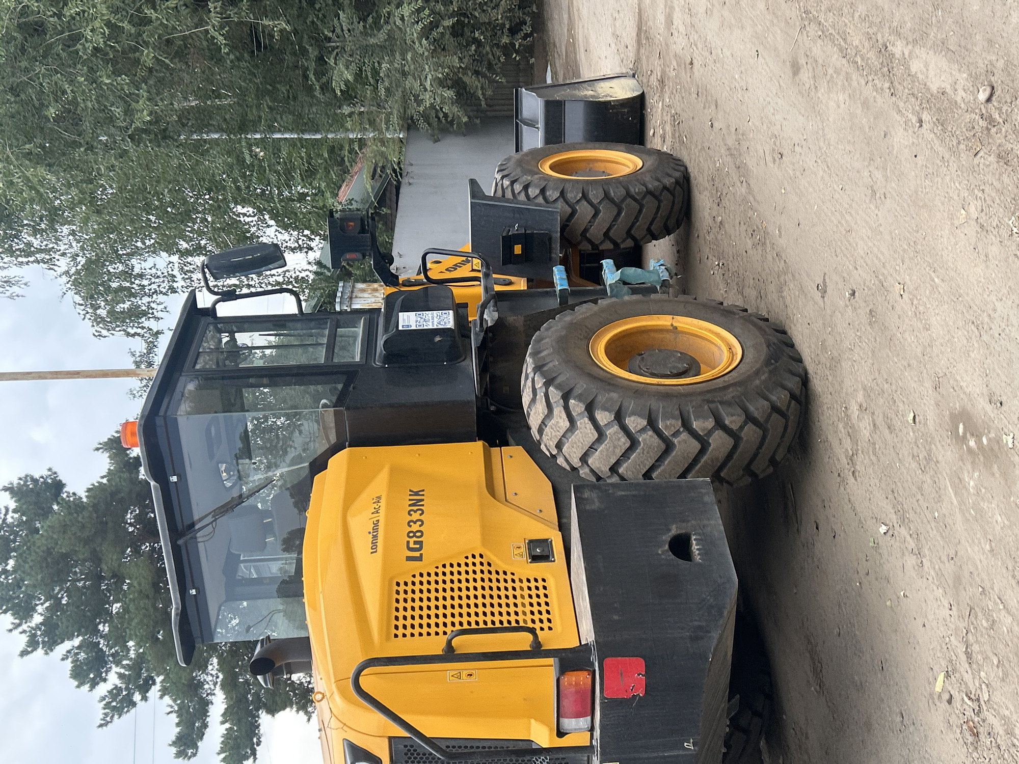 A yellow and black wheel loader, identified by the "LONKING Ac-Ai LG833NK" marking on its side, is parked outdoors on a dusty, unpaved surface, likely a construction site or industrial area. The loader is positioned at an angle, with its front facing towards the right of the frame. Large, rugged tires with deep treads are visible. The machine has a closed cab with tinted windows and a windshield wiper. An orange flashing light is mounted on top of the cab. Behind the loader, there is a grey concrete wall and dense green foliage, suggesting an outdoor environment with vegetation. The sky is overcast. There are no people visible in the image. The location is described as Almaty, Kazakhstan.