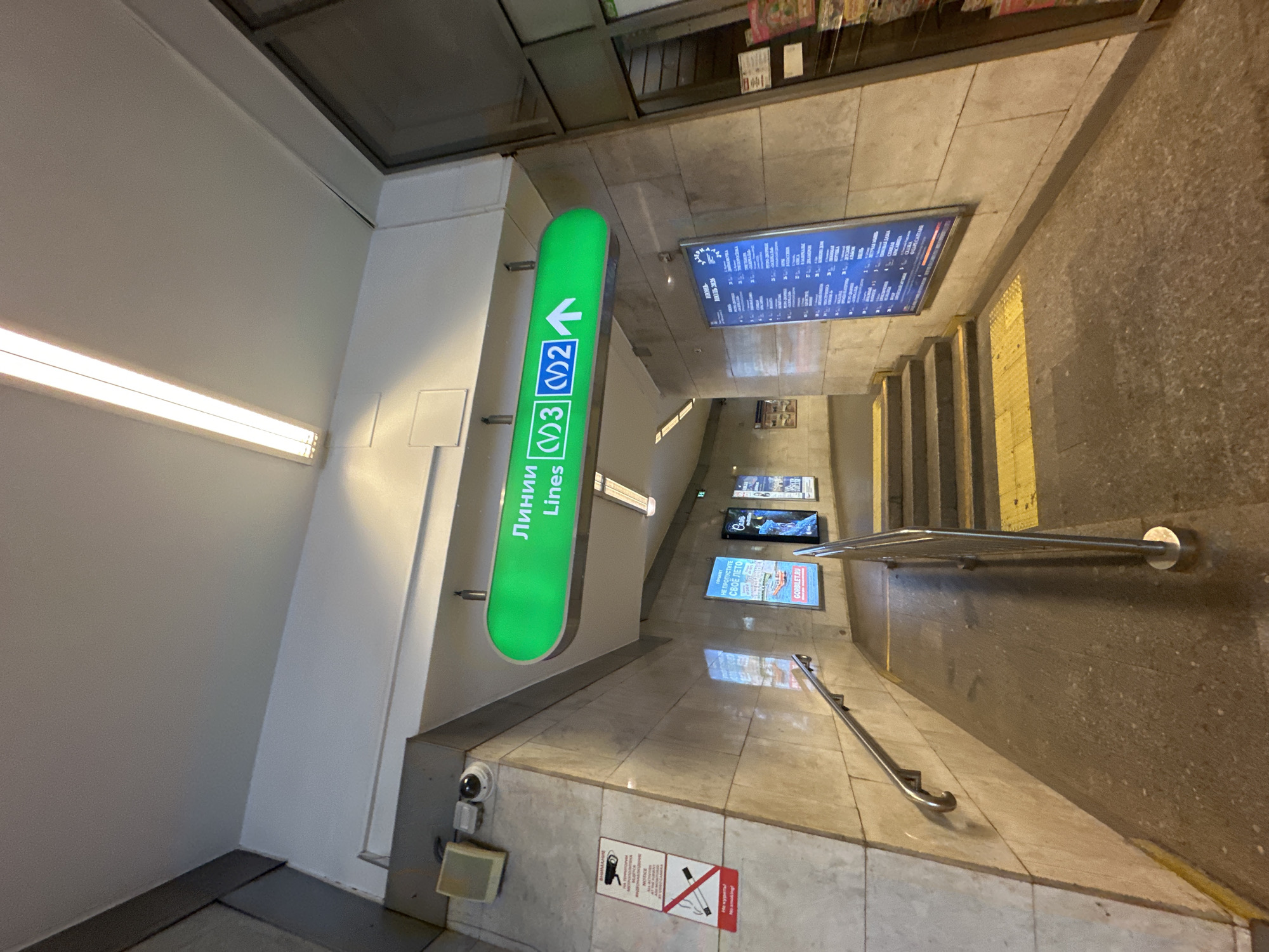 The image shows an underground passageway in a city. A large green sign overhead reads "Линии Lines" and indicates directions to lines M3 and M2 with an arrow pointing right. The walls are clad in beige marble with a textured finish. A metal handrail runs along the left wall, and a set of stairs with yellow tread markings is visible ahead. To the left, there are signs about CCTV surveillance and no smoking. On the right, a large digital display board shows a schedule or map, and a rack of magazines is visible. The lighting is provided by fluorescent tubes on the ceiling. The environment appears to be a public transit station, likely a metro station, given the signage. There are no people visible in the image.