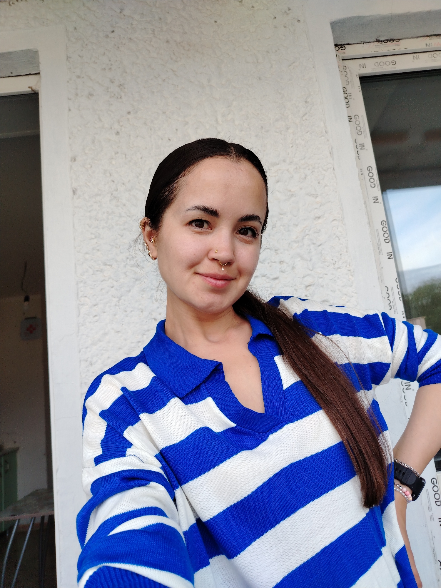 A young woman is centered in the frame. She has dark hair pulled back in a low ponytail, light skin, and is wearing a blue and white striped sweater. She has multiple ear piercings on her left ear and a nose ring. The background consists of a textured white wall and a white-framed window with the words "GOOD IN" printed repeatedly around the frame. A partial view of a green object and metal railing are visible through the window. The photograph is well-lit and appears to be taken indoors. The location is likely in Noviy, Russia, based on the prompt.