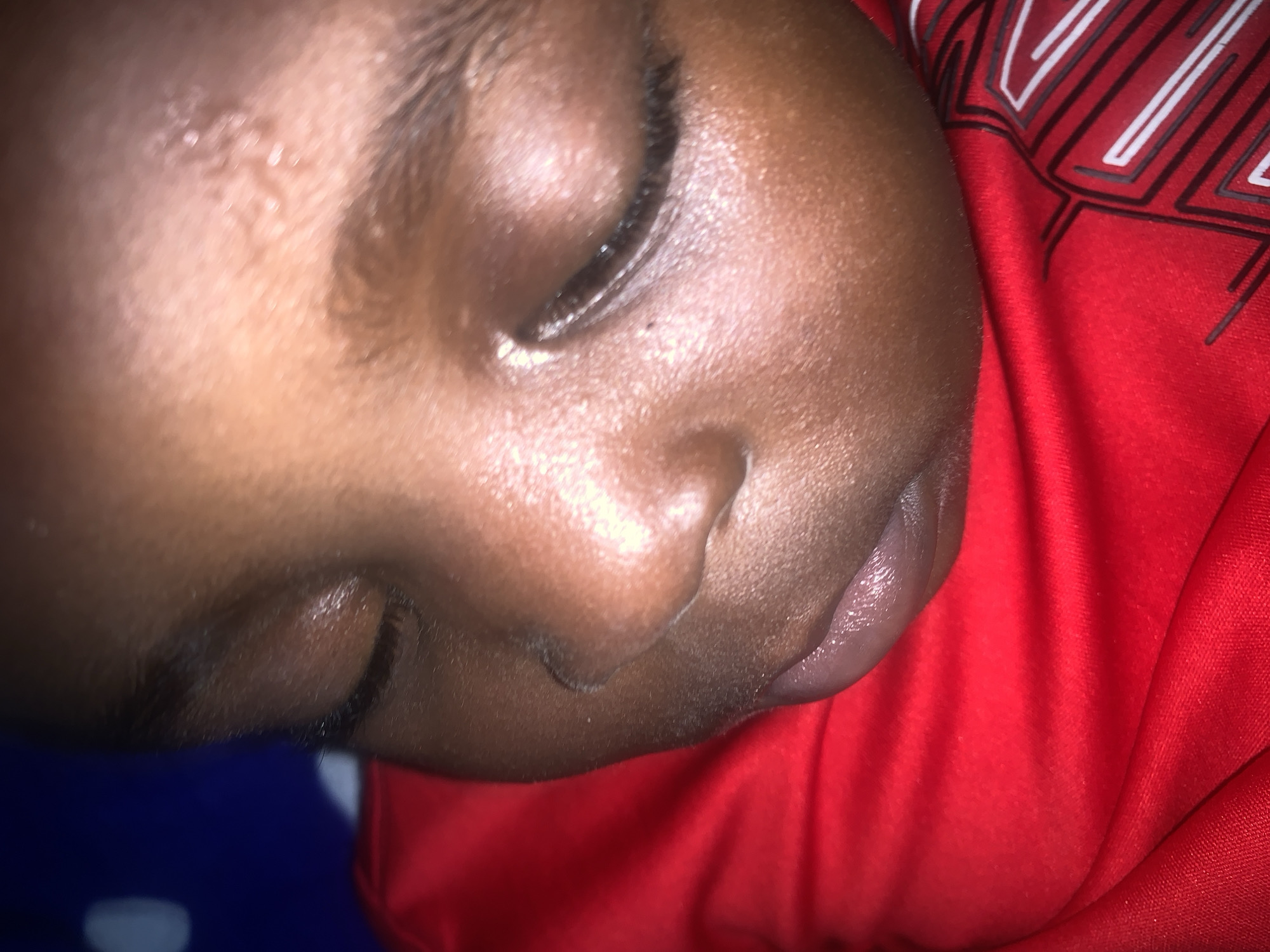 This is a close-up, low-angle, indoor, eye-level, medium shot of the face of a young child who appears to be sleeping. The child has dark skin and dark hair. The child is wearing a red shirt with a graphic design on the right side of the frame. The background is blurred and appears to be dark blue with some lighter circular shapes. The lighting is dim, with some highlights on the child's forehead and cheek. The image is slightly out of focus, particularly around the edges.