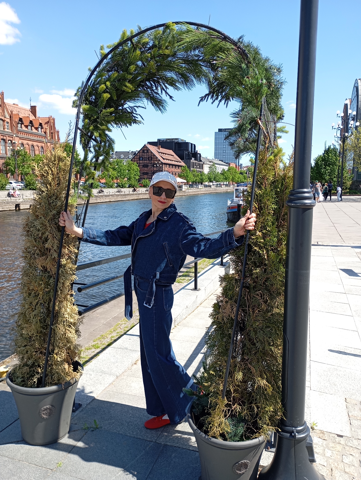A full shot captures a woman standing on a sidewalk in Bydgoszcz, Poland, positioned beneath an archway of greenery. She wears a denim jacket and matching trousers, a white cap, sunglasses, and red shoes. The archway, set in pots, frames her against a backdrop of the Brda River and historic brick buildings. A black lamppost stands to her right. The sky is clear and blue. Other pedestrians are visible in the background.