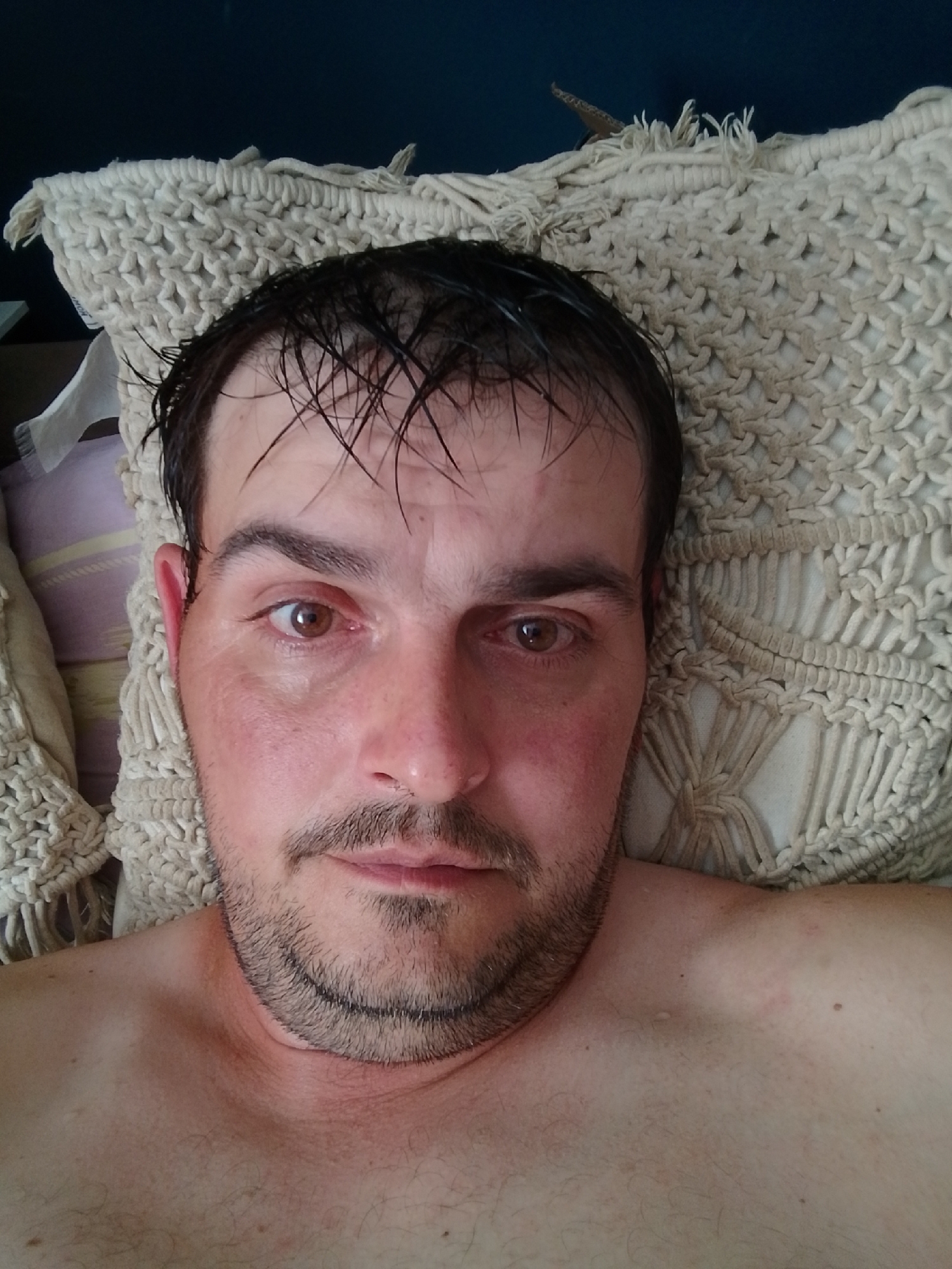 A close-up, selfie-style photograph features a man lying down, with his head resting on a beige, crocheted pillow. The man has short, dark, wet hair, brown eyes, and a light beard and mustache. His skin appears flushed on his cheeks and neck. He is wearing a light-colored garment that is mostly out of frame, revealing his bare shoulder and chest. The background consists of a dark blue wall and a patterned fabric behind the pillow. The scene appears to be indoors.